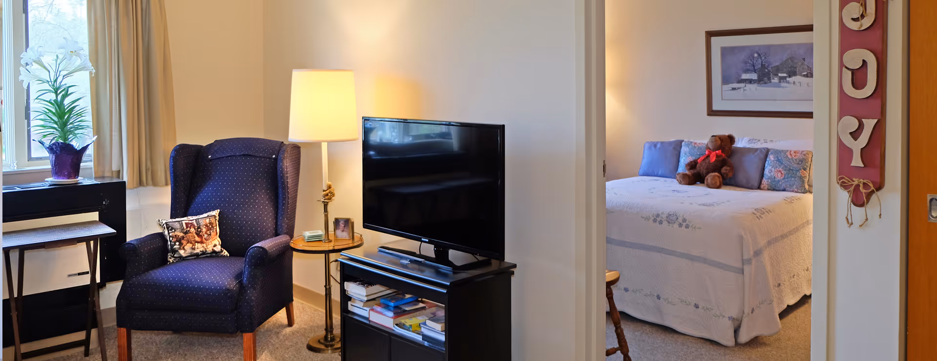 A cozy living area with a dark blue armchair, a small round side table with a lamp and a framed photo, a flat-screen TV on a black stand with books underneath, and a window with beige curtains and a potted white flower. Through an open doorway, a bedroom is visible with a bed covered in a white quilt, blue and floral pillows, a teddy bear with a red bow, a framed winter landscape painting on the wall, and a wooden chair partially visible.