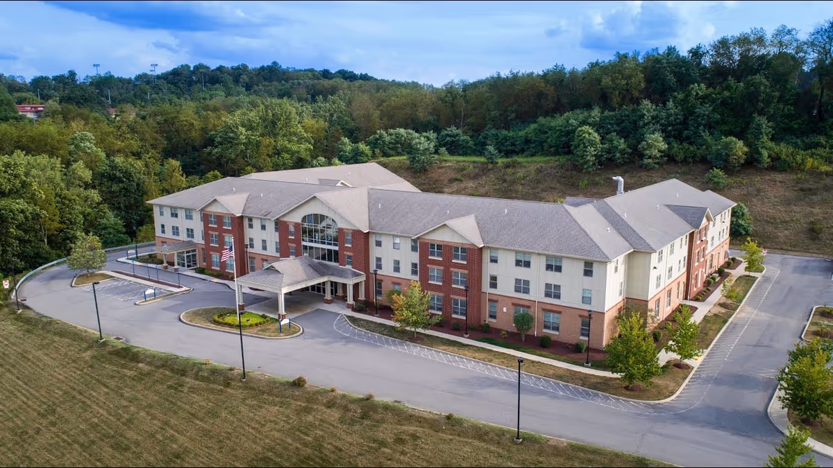 Aerial view of a three-story senior living facility building with a covered entrance, surrounded by a driveway and parking areas. The building has a mix of brick and light-colored siding exterior, with multiple windows and a large central window above the entrance. The facility is set against a backdrop of dense green trees and a partly cloudy sky.