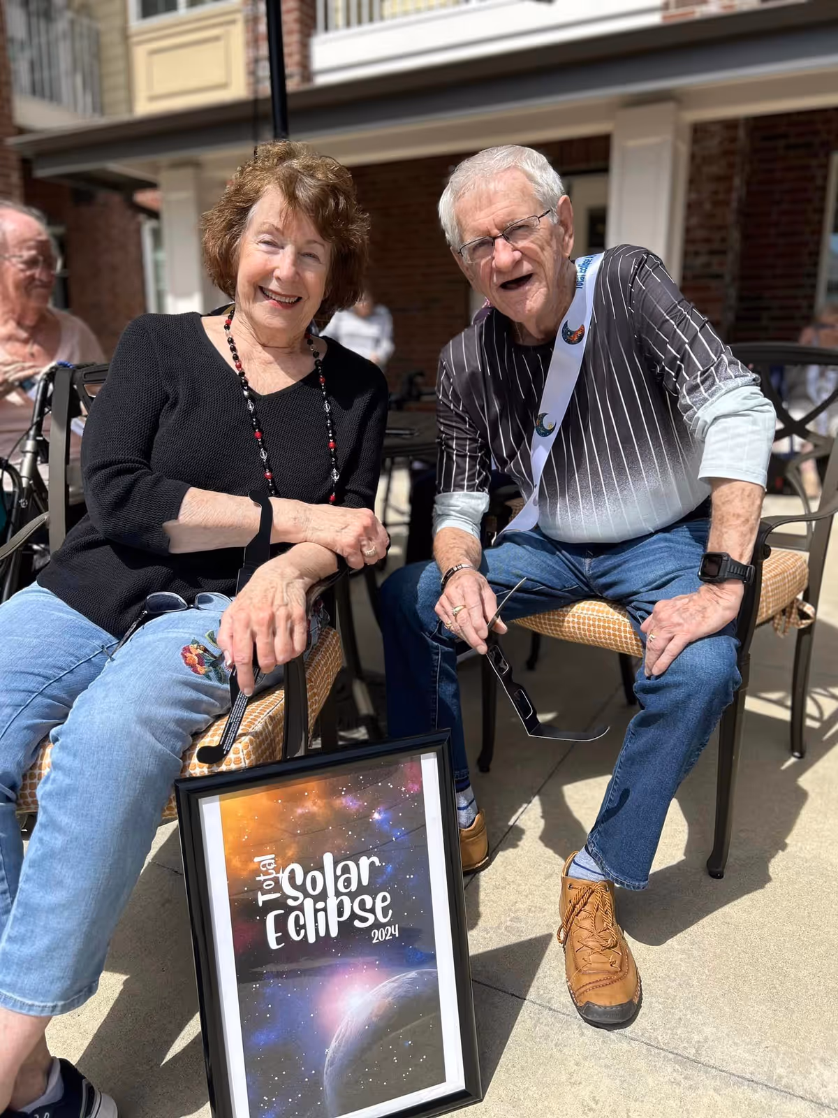 Two elderly people sitting outdoors on chairs, smiling at the camera. The woman on the left is wearing a black top and light blue jeans, while the man on the right is wearing a black and white striped shirt with blue jeans and brown shoes. They are holding a framed poster that reads 'Total Solar Eclipse 2024'. The setting appears to be a patio area with a brick building in the background.