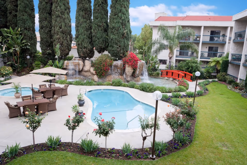 Outdoor area of Arcadia Gardens Retirement Hotel featuring a swimming pool, hot tub, patio with tables and chairs under umbrellas, landscaped garden with flowers and bushes, tall trees, a waterfall feature with rocks, a red wooden bridge, and a multi-story building with balconies in the background under a partly cloudy sky.