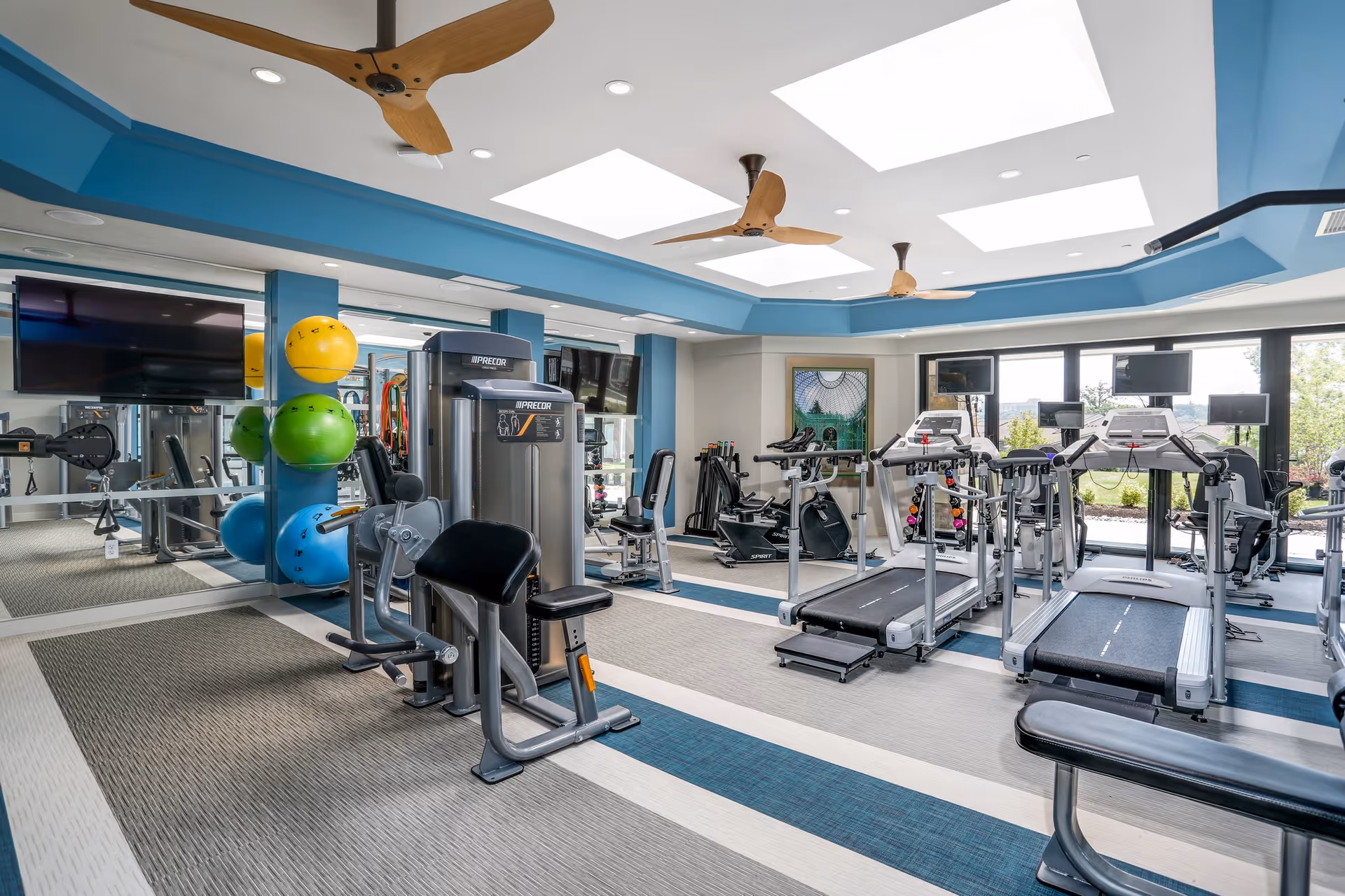 Bright fitness room with treadmills, weight machines, colorful exercise balls, and ceiling fans.