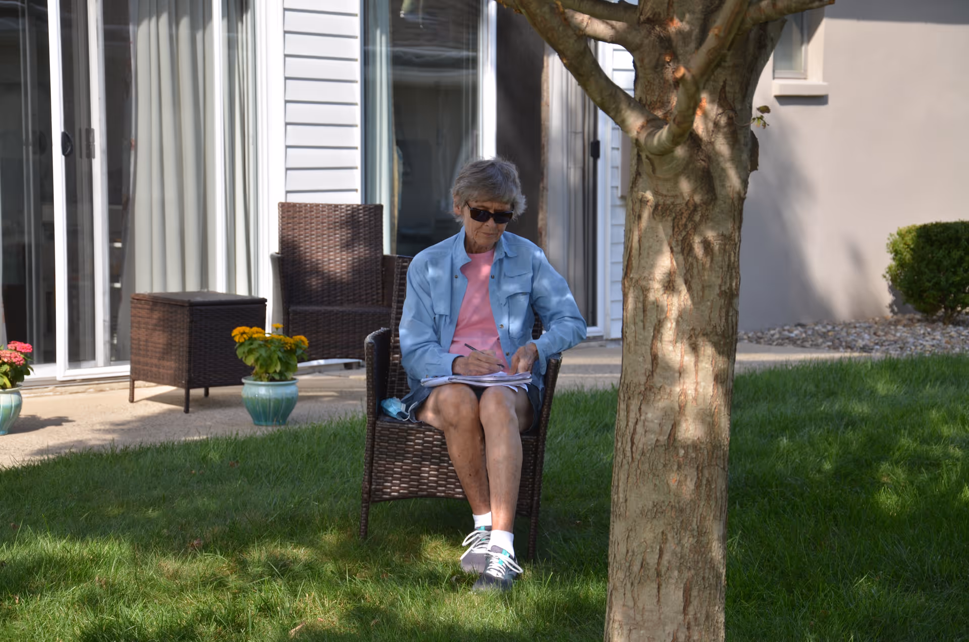 An elderly woman wearing sunglasses, a light blue jacket, pink shirt, and sneakers is sitting on a wicker chair on a grassy lawn near a tree. She is writing or drawing on a notepad. Behind her is a building with sliding glass doors, potted flowers, and outdoor wicker furniture.