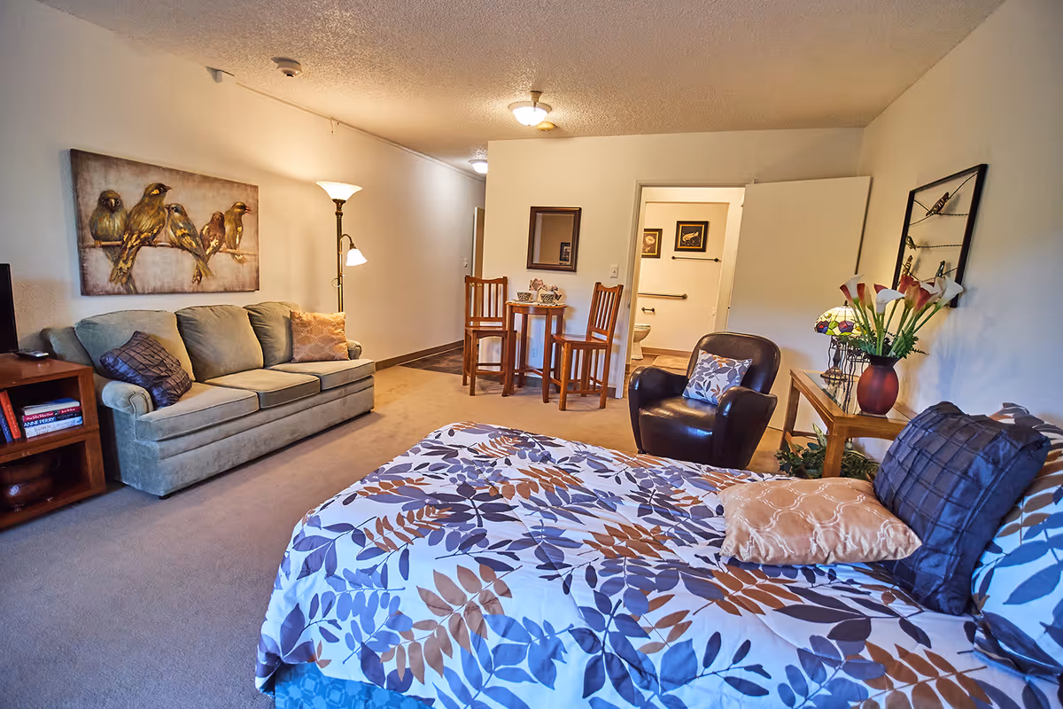 A senior living facility room featuring a bed with a floral patterned bedspread, a green sofa with pillows, a wooden side table with books, a floor lamp, a small round table with two wooden chairs, a leather armchair with a pillow, and a wooden console table with a vase of flowers. A bathroom door is open in the background, showing grab bars and framed pictures on the wall.
