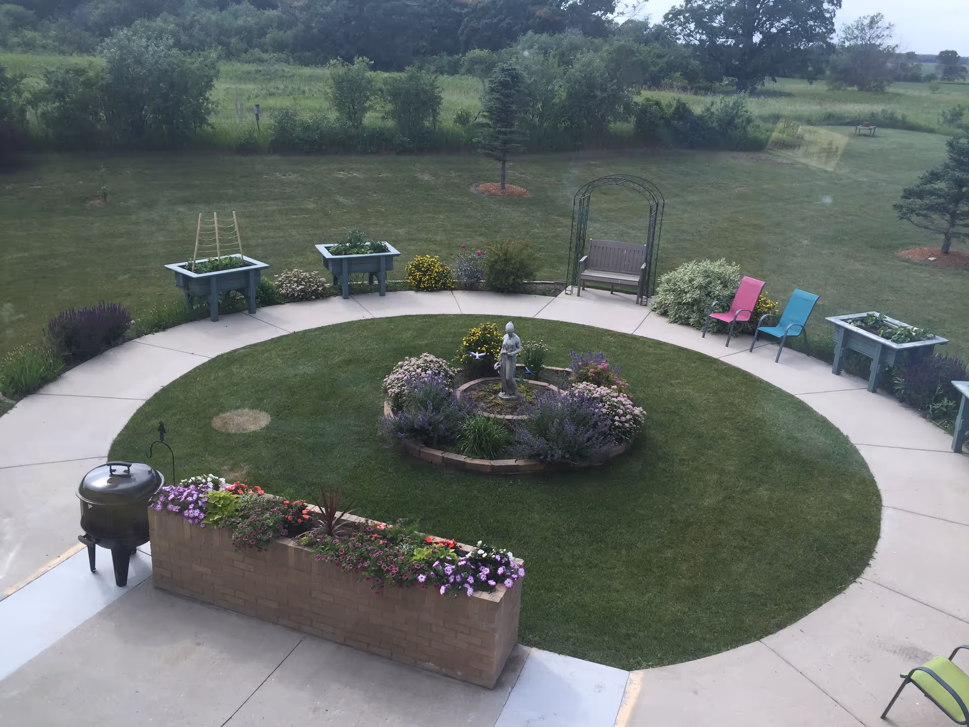Circular outdoor courtyard with a central flowerbed and statue, surrounded by a concrete walkway, raised planters, benches and colorful chairs.