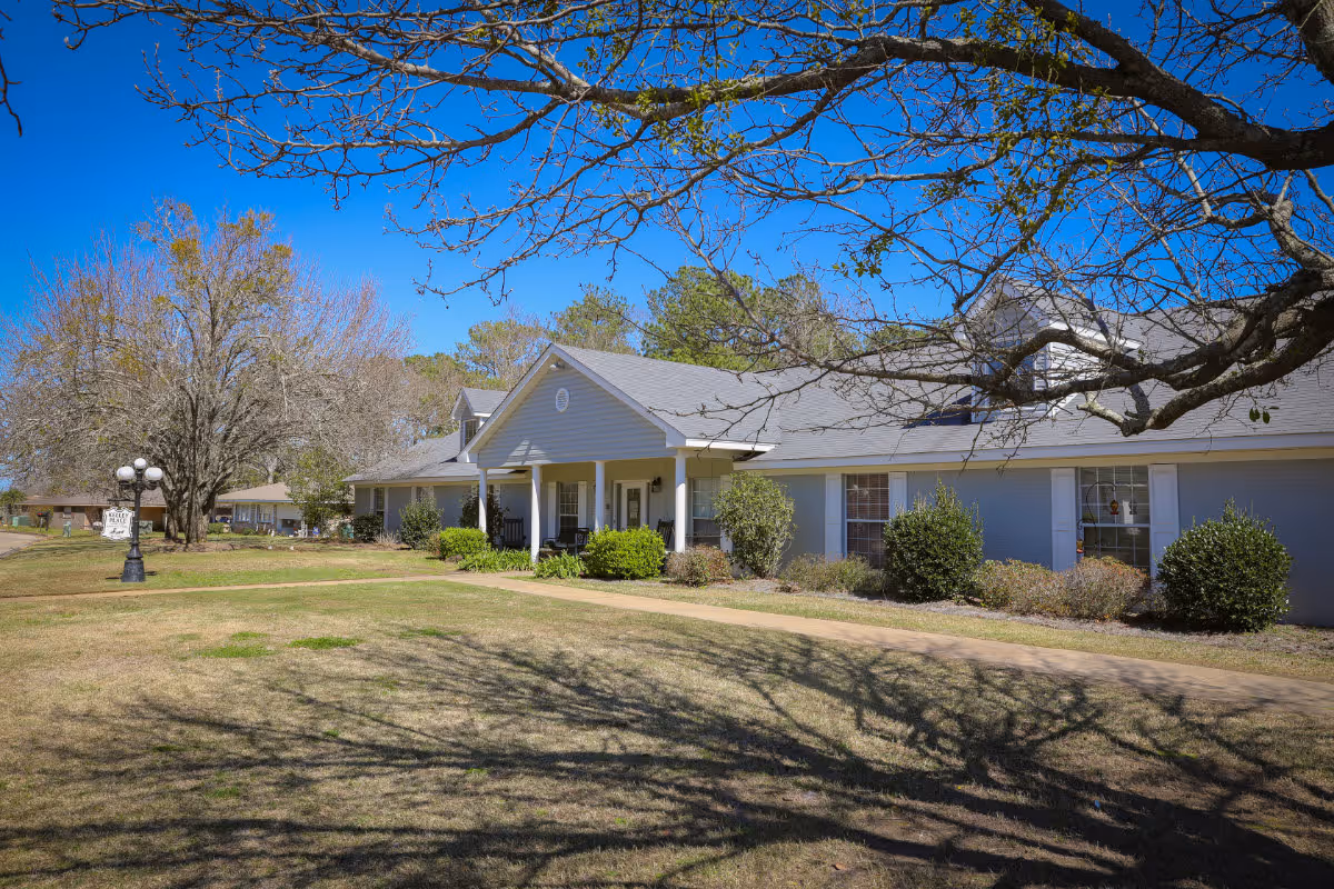 Exterior view of a single-story building with a gray roof and light blue walls, surrounded by bushes and trees with some leaves. A paved walkway leads to the entrance with white columns and a small porch. The sky is clear and blue.