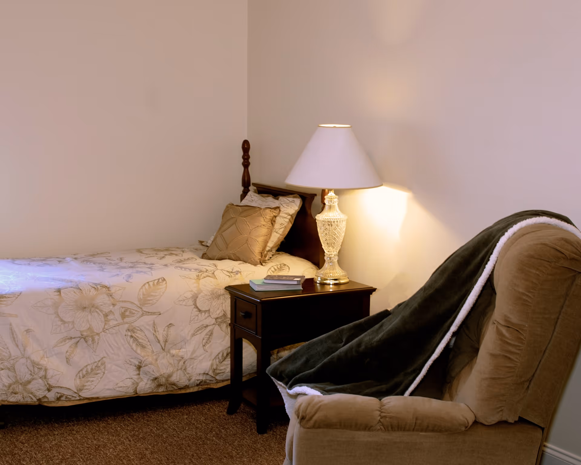 A cozy bedroom with a single bed covered in a floral bedspread and a decorative pillow. Next to the bed is a dark wooden nightstand with two books and a glass table lamp with a white lampshade. In front of the nightstand is a cushioned armchair draped with a dark green blanket with a white edge.
