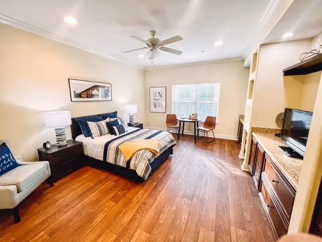 A well-lit bedroom with wooden flooring featuring a neatly made bed with striped bedding and multiple pillows. There are two bedside tables with lamps, a ceiling fan, a small table with two chairs near a window, a comfortable armchair with a pillow, and a built-in cabinet with a TV on the right side.