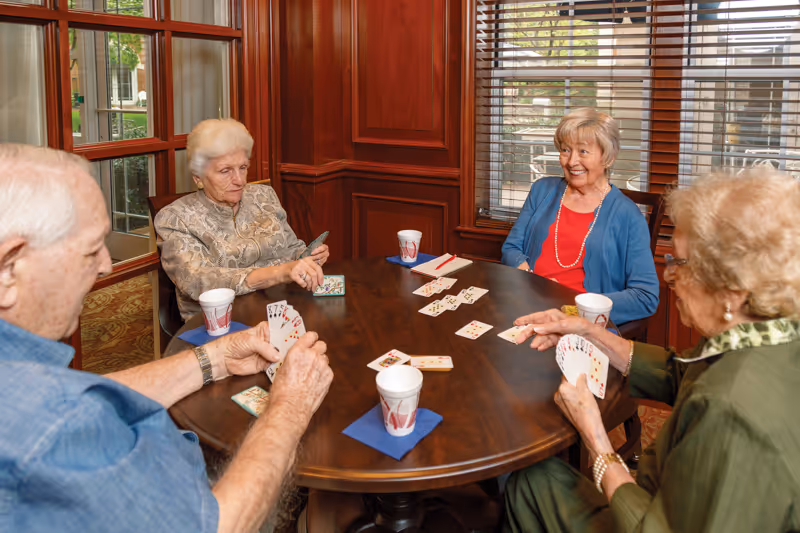 Four elderly people sitting around a wooden round table playing a card game in a room with wooden paneling and large windows with blinds. Each person has a paper cup on a blue napkin in front of them.