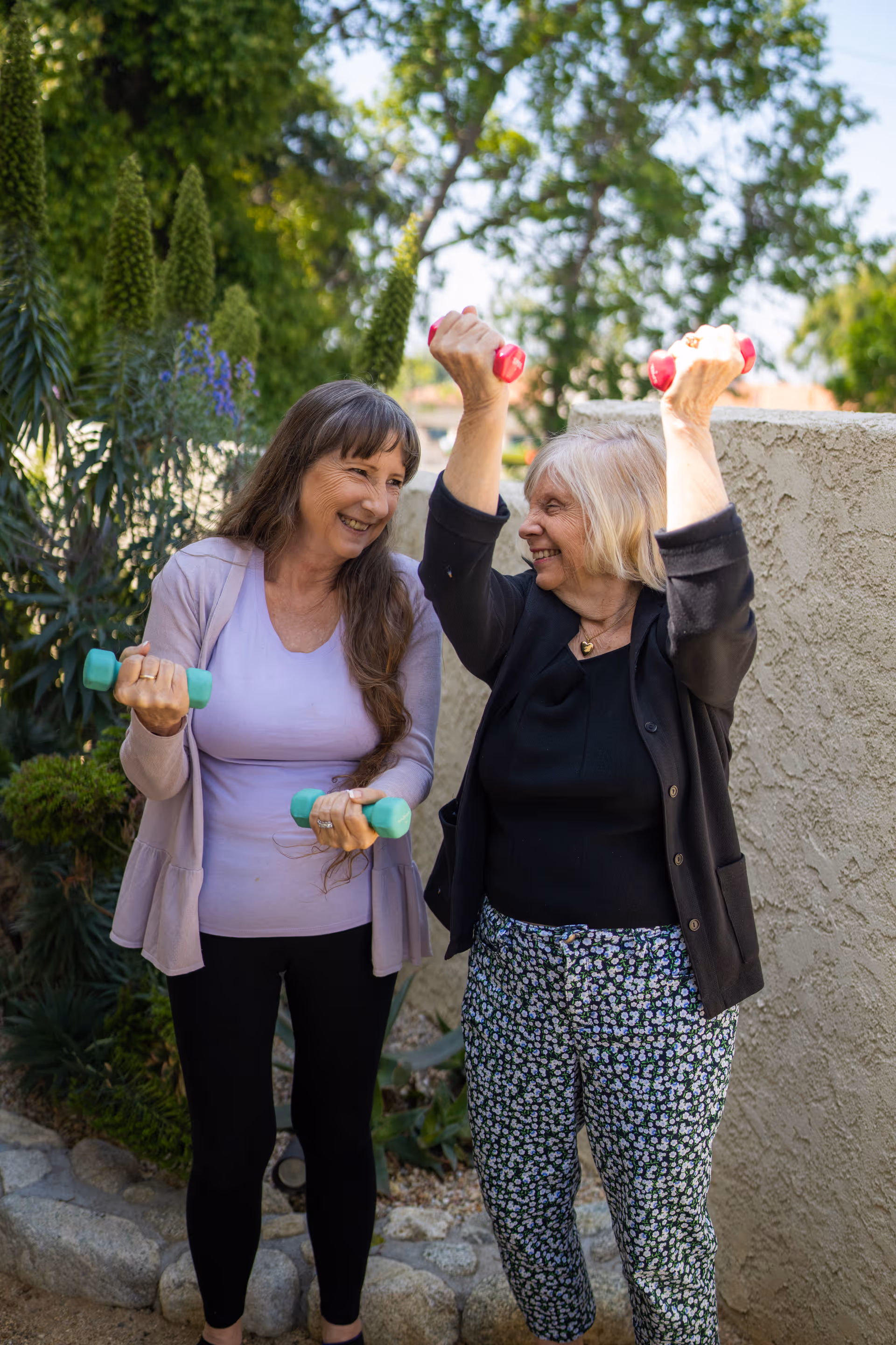 Two elderly women smiling and exercising outdoors with small dumbbells, surrounded by greenery and a stone wall in the background.