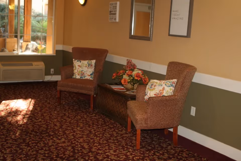 A cozy seating area with two upholstered armchairs featuring floral patterned cushions, a wooden chest serving as a table with a flower arrangement on top, a window letting in natural light, and framed wall art including a mirror and a motivational quote that says 'MAKE TODAY AMAZING'.