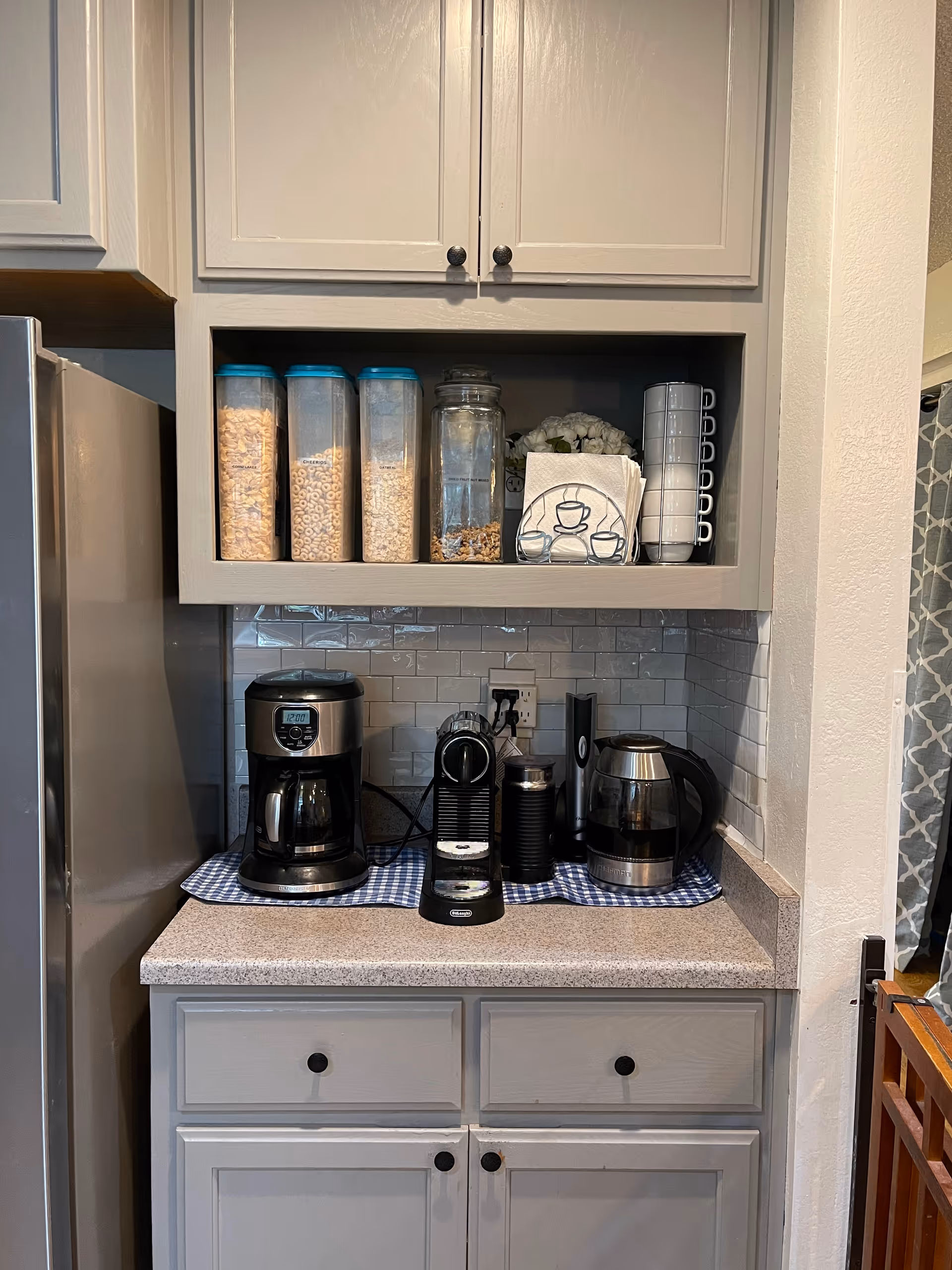 A kitchen counter area with a coffee maker, espresso machine, electric kettle, and a milk frother placed on a blue and white checkered cloth. Above the counter, there are three cereal containers, a jar with granola, a stack of white cups, and napkins with a coffee cup design. The cabinets and drawers are painted light gray, and the backsplash is made of white subway tiles.
