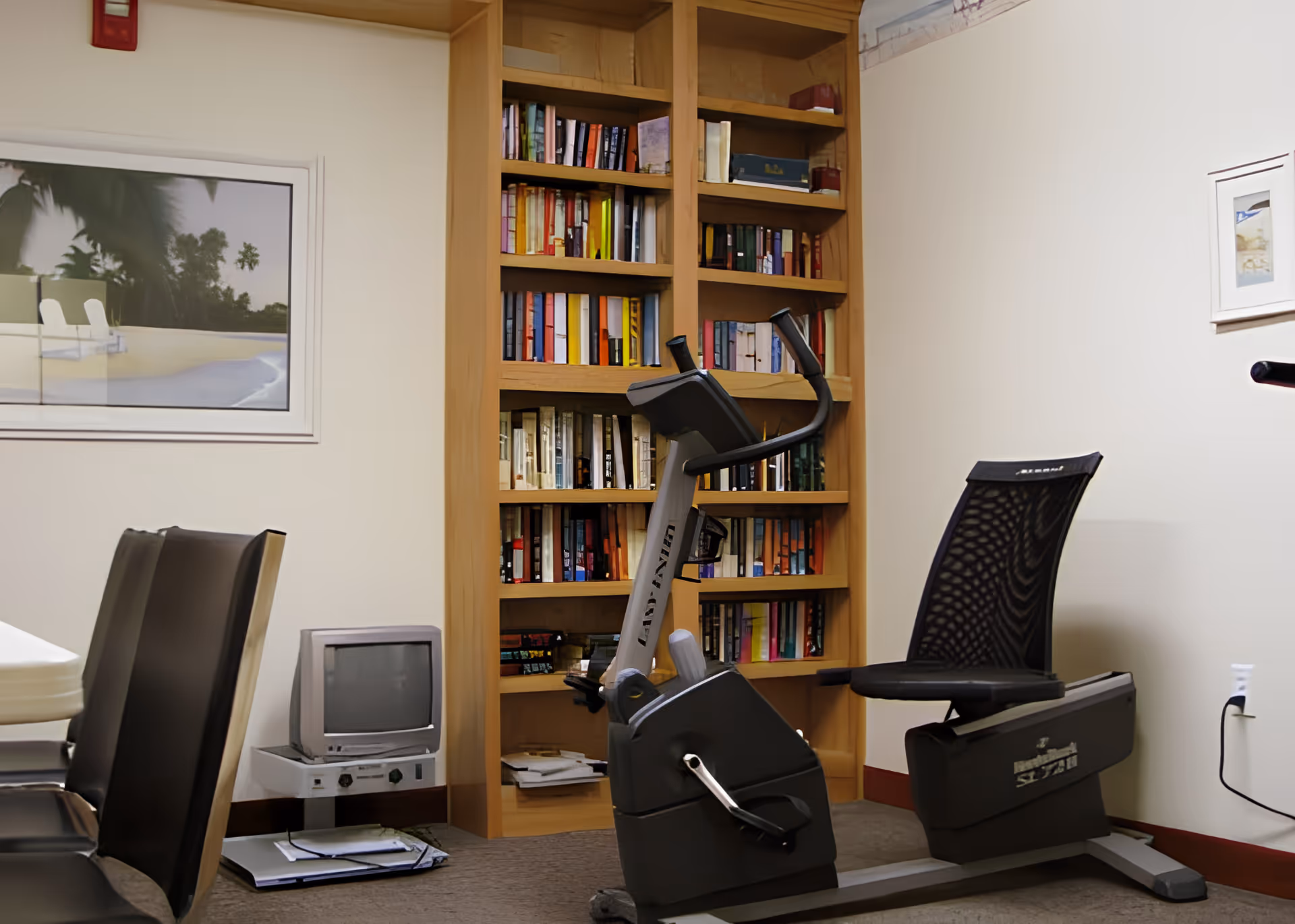 A room with a recumbent exercise bike in front of a wooden bookshelf filled with books. To the left, there are black chairs and a small old-style television on a stand. The walls are light-colored with framed pictures hanging on them.