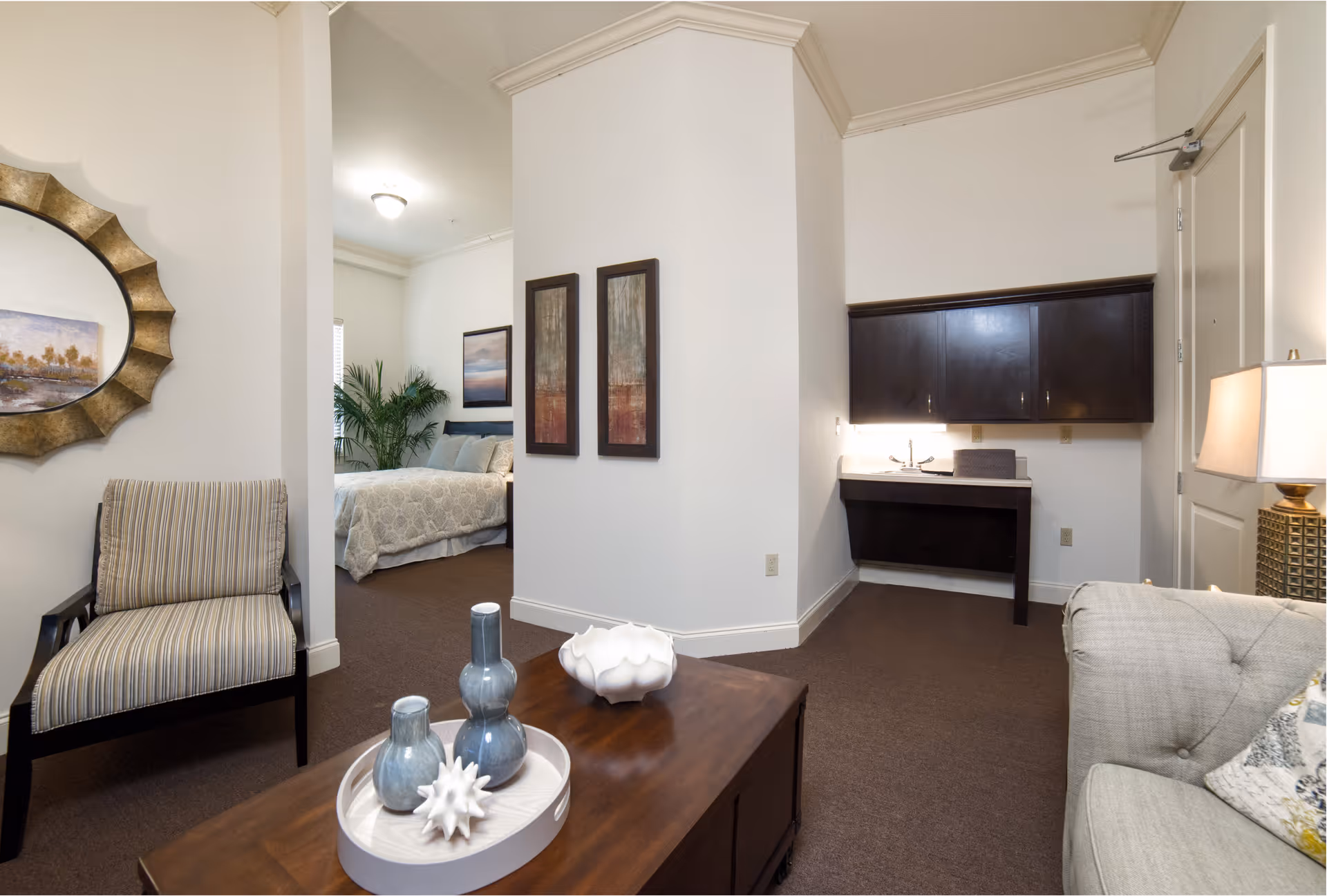 Interior view of a senior living facility suite showing a living area with a striped armchair, a wooden coffee table with decorative vases, a beige couch with a pillow, and a lamp. In the background, there is a bedroom with a bed, artwork on the walls, and a large plant. There is also a small desk area with dark cabinets mounted on the wall.
