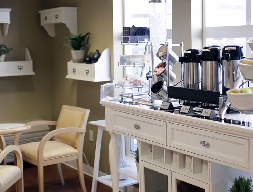 A cozy interior corner of a senior living facility with a white wooden sideboard holding coffee dispensers labeled Coffee, Hot Water, and Decaf, along with cups and condiments. Two cushioned wooden chairs with yellow upholstery are positioned nearby, and decorative shelves with plants and small ornaments are mounted on the olive-green wall.