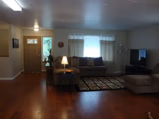 Spacious living room with hardwood floors, a sofa and ottoman by a patterned rug, TV on a stand, and an entry door at left.