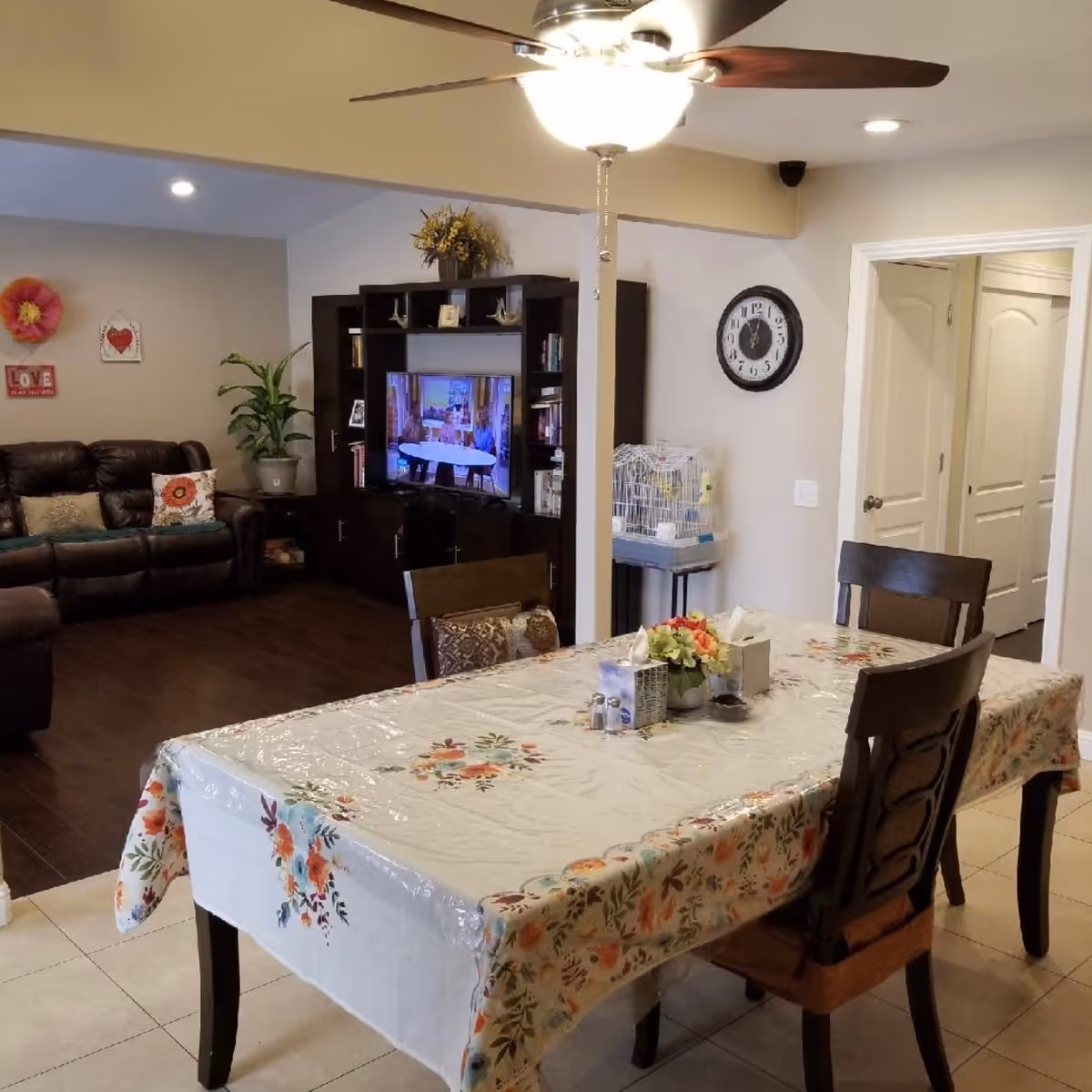 Dining table with a floral tablecloth and chairs in an open-plan room showing a living area with a TV, wall clock, and ceiling fan.