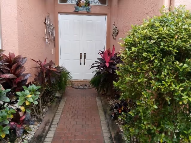 A narrow brick pathway lined with various green and red-leaved plants on both sides leading to a set of white double doors on a peach-colored building.