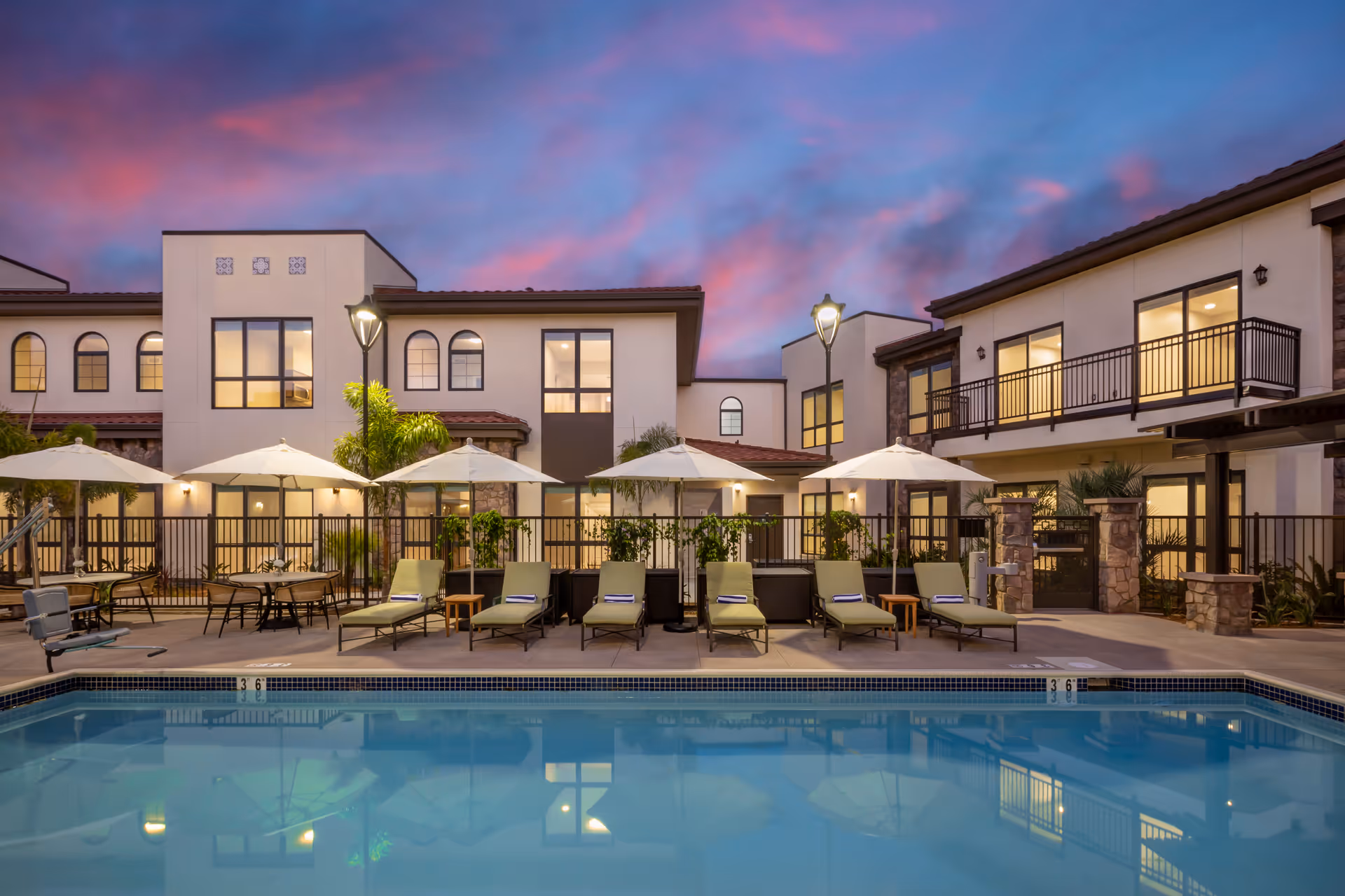 Outdoor swimming pool and patio area with lounge chairs, umbrellas, and a two-story building lit at dusk.