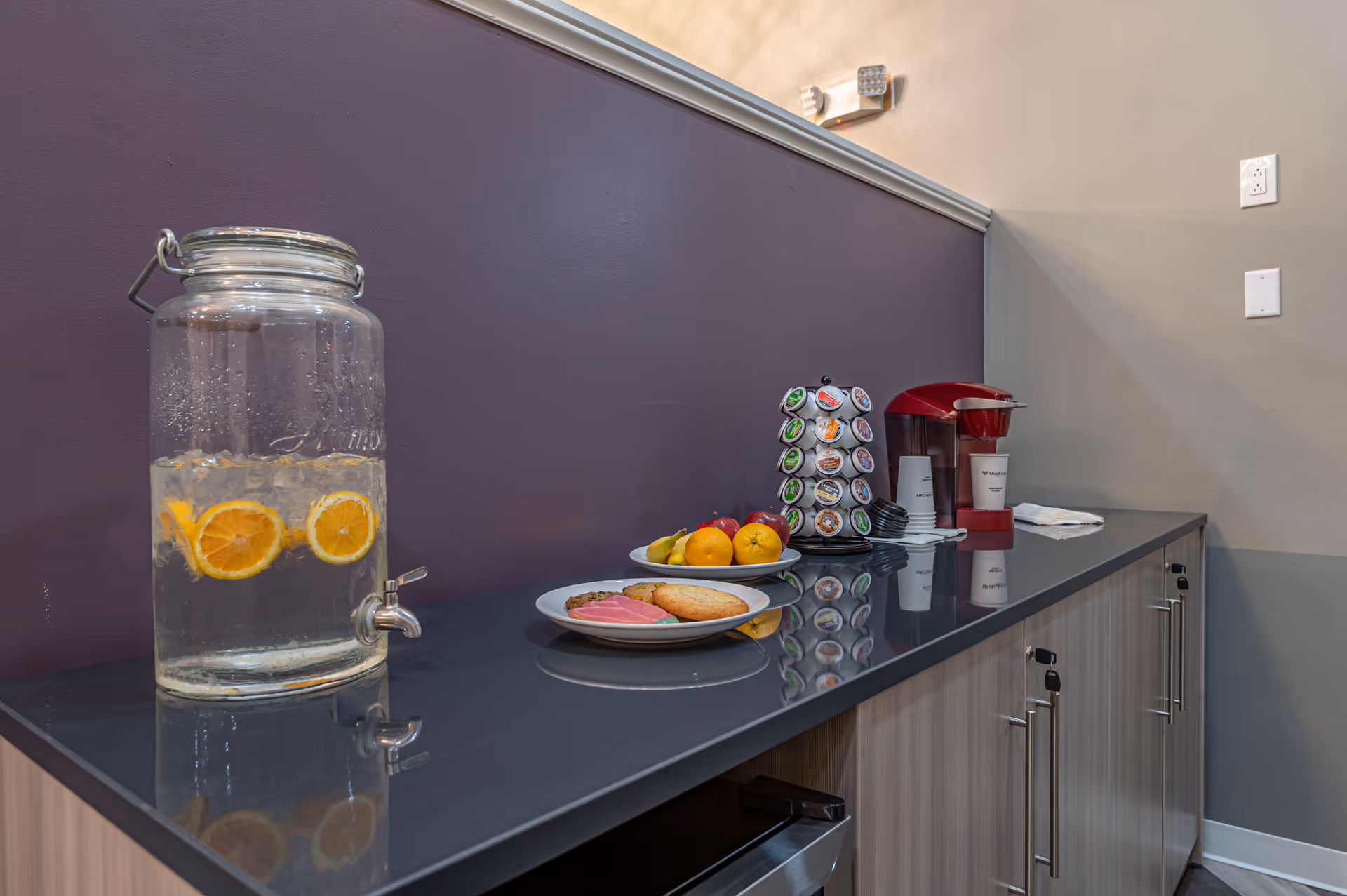 A countertop with a glass dispenser filled with water and orange slices, two plates with cookies and fruit, a rotating rack with coffee pods, a red coffee maker, and some disposable cups and napkins against a purple and beige wall.
