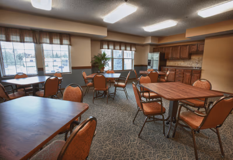 A dining room with several wooden tables and cushioned chairs arranged around them. The room has large windows with blinds, a carpeted floor, and a kitchenette area with wooden cabinets and a stainless steel refrigerator in the background.