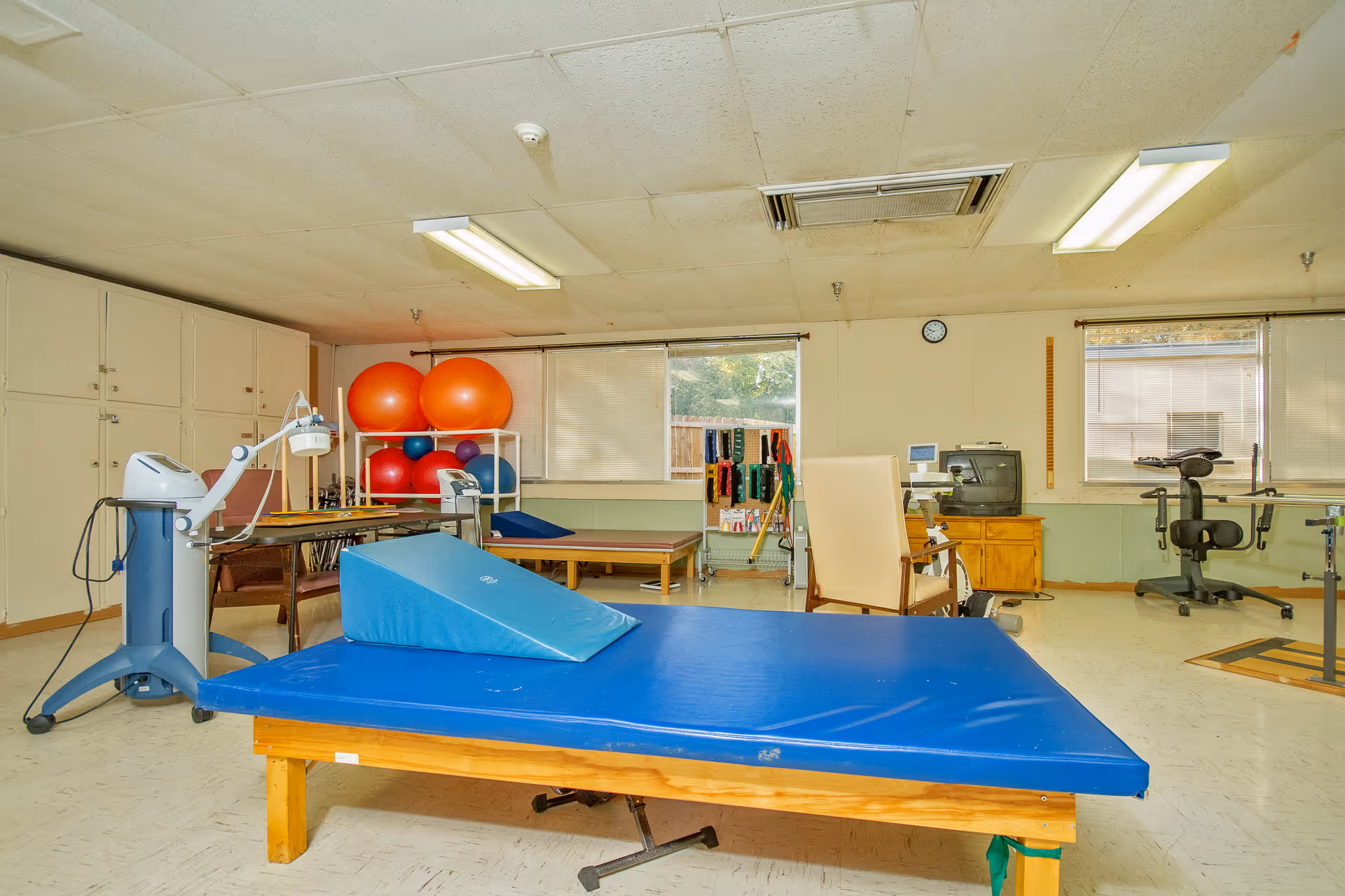 A physical therapy room with a large blue padded therapy table in the center, exercise balls stacked on a rack, various therapy equipment including a stationary exercise bike, and a chair. The room has two windows with blinds, a clock on the wall, and fluorescent ceiling lights.