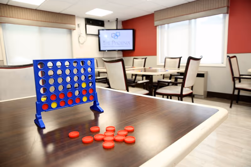 A game of Connect Four set up on a wooden table in a well-lit room with several chairs and tables. A television mounted on the wall displays the logo and name 'The Phoenix Rehabilitation & Nursing Center'. The room has large windows with blinds and red and white walls.