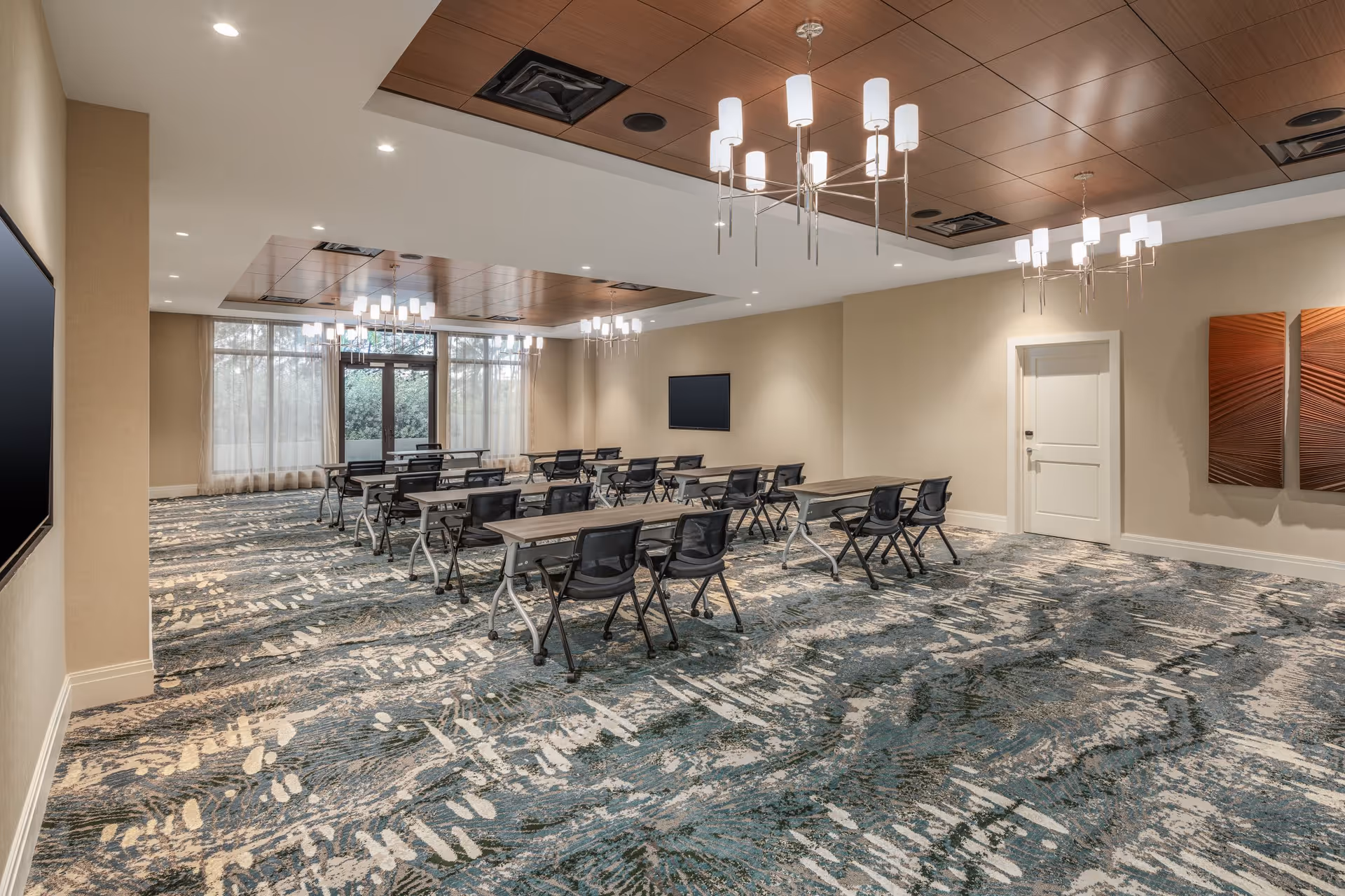 A spacious, well-lit conference or meeting room with multiple rows of tables and black chairs arranged facing a wall-mounted flat screen. The room features modern chandeliers, a patterned carpet in shades of blue and beige, beige walls, and large windows with sheer curtains allowing natural light to enter.