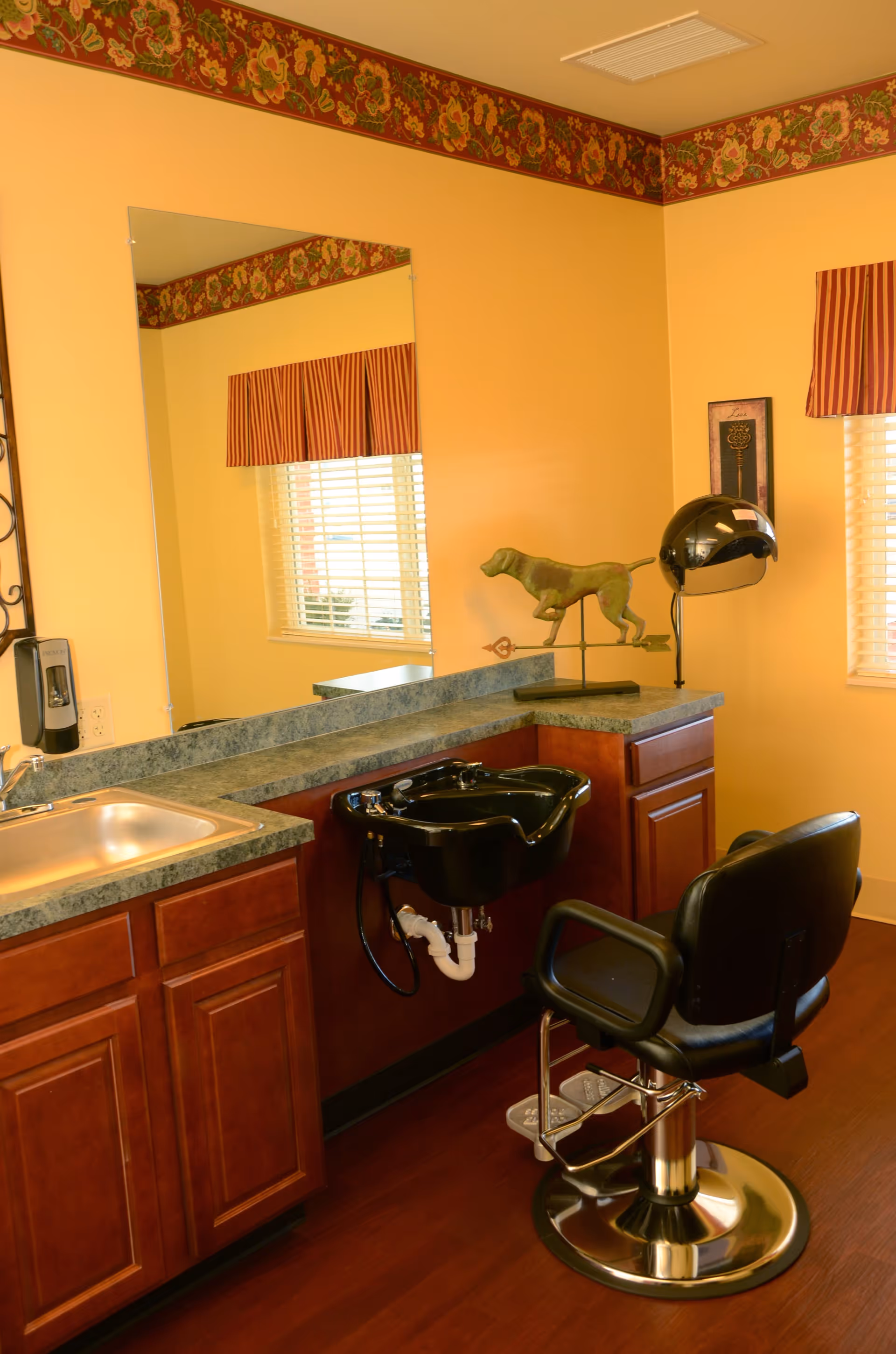 Interior view of a hair salon area within a facility, featuring a black salon chair in front of a black hair washing sink. The countertop is greenish with wooden cabinets below. There is a large mirror on the wall, a window with striped valance curtains, and a decorative dog sculpture on the counter. The walls are painted yellow with a floral border near the ceiling.