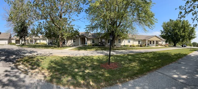 Wide exterior view of a single-story senior living facility building with beige walls and a brown roof, surrounded by green grass and several trees under a clear blue sky.