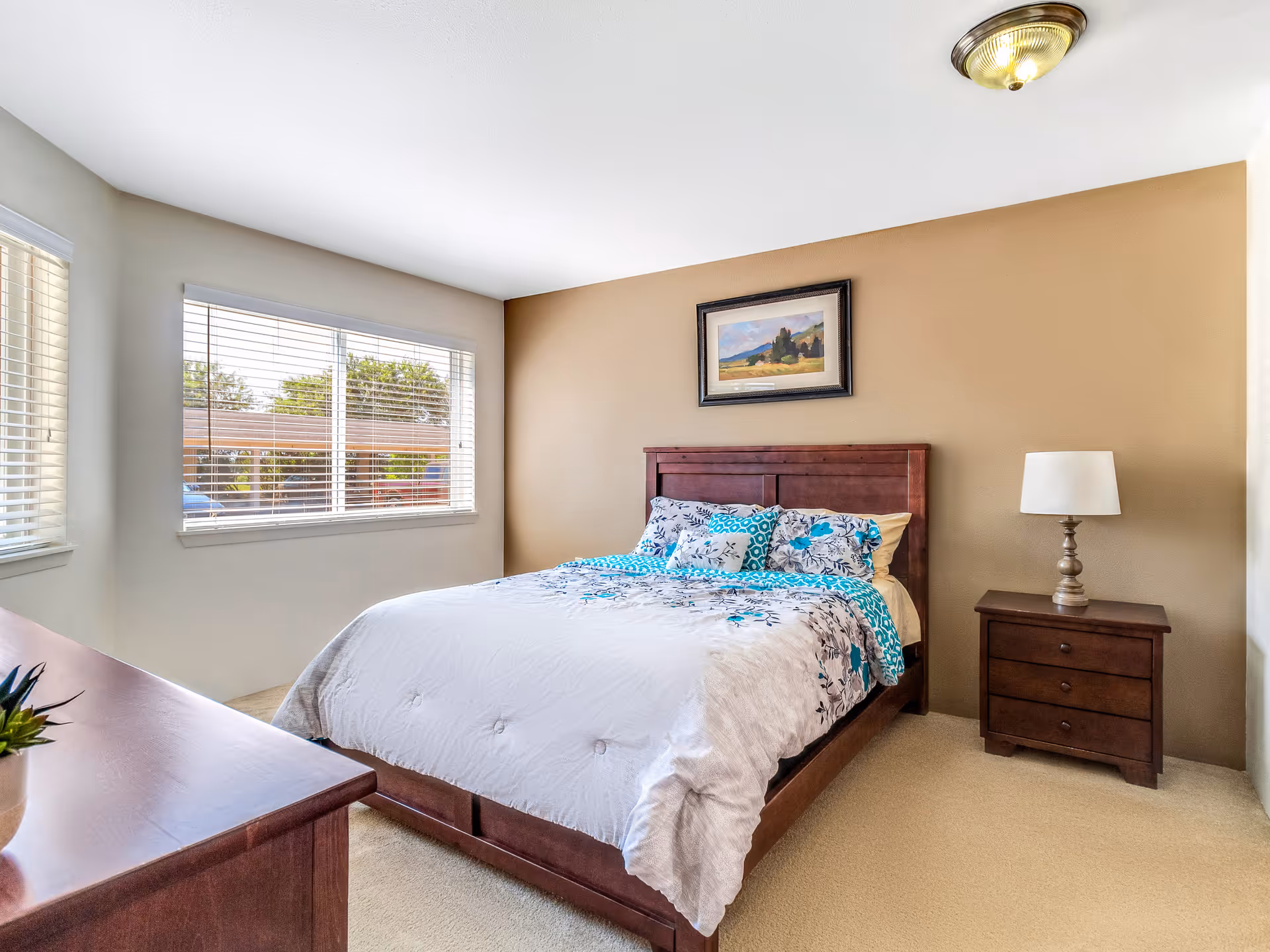 A bright bedroom with a large window with blinds, a wooden bed with floral bedding in white, blue, and gray tones, a matching wooden nightstand with a lamp, a wooden dresser with a small plant, beige walls, and a framed landscape painting above the bed.