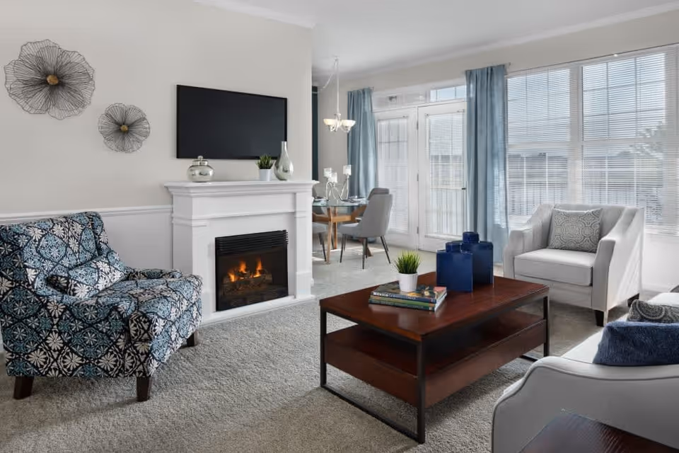 A bright and cozy living room featuring a patterned blue armchair, a white armchair with a decorative pillow, a wooden coffee table with books and blue vases, a white fireplace with a flat-screen TV mounted above it, and large windows with blue curtains letting in natural light. In the background, a dining area with a round glass table and gray chairs is visible.