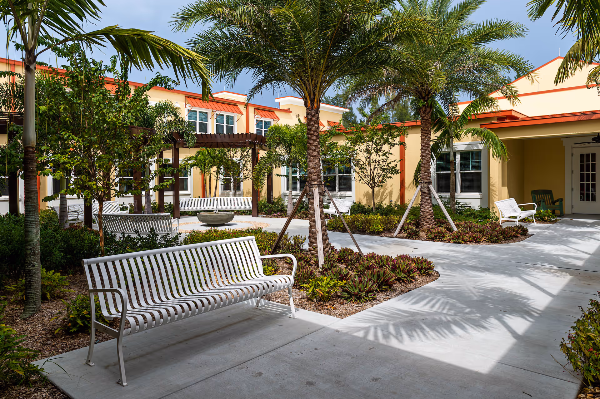 Outdoor courtyard area at The Gallery at Cape Coral featuring white metal benches, palm trees, various plants, and a pergola structure with a fire pit in the center. The building surrounding the courtyard has yellow walls with red trim and multiple windows.