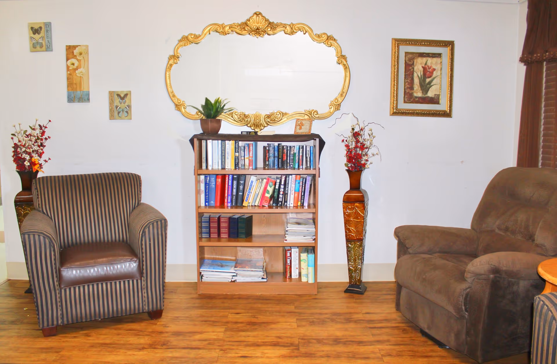 A cozy living room area with a striped armchair on the left and a brown recliner on the right. Between them is a wooden bookshelf filled with books and magazines. Above the bookshelf is an ornate gold-framed mirror. On either side of the bookshelf are tall decorative vases with red and white flowers. The walls are decorated with small framed pictures and a floral painting. The floor is wooden.