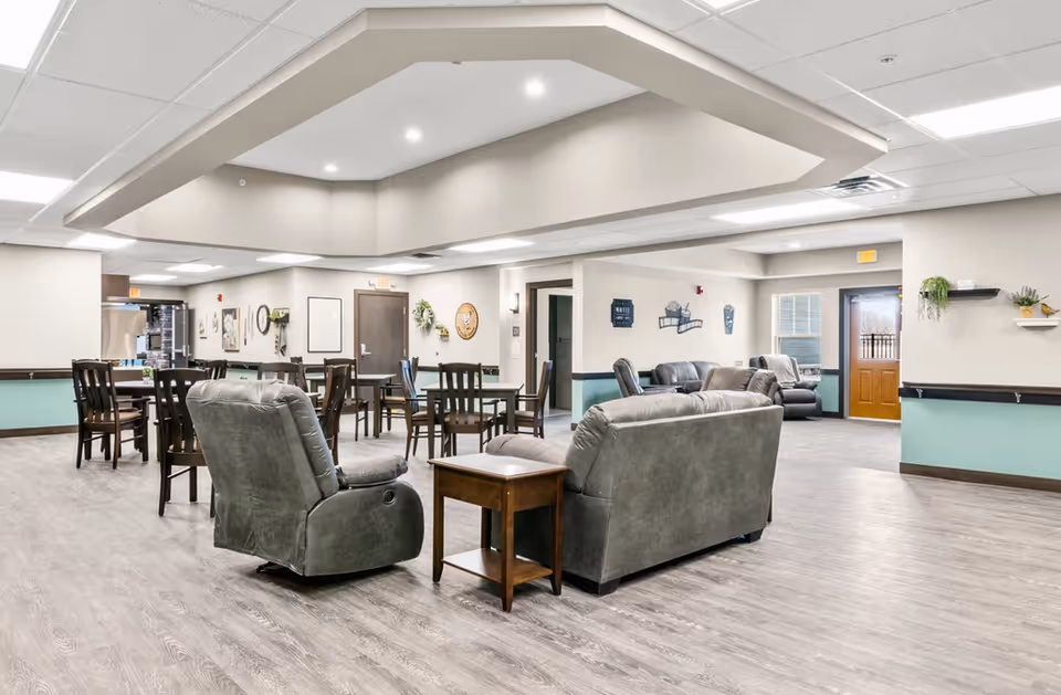 A spacious senior living common area with multiple seating options including gray leather sofas and armchairs, wooden tables and chairs, light wood flooring, and soft neutral wall colors with teal accents. The room has a modern ceiling design with recessed lighting and a wooden door leading outside.
