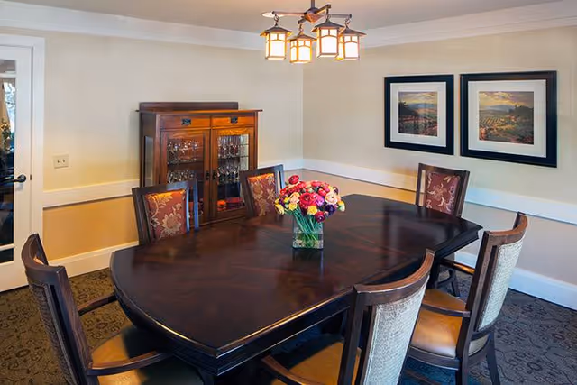 A dining room with a large dark wooden table surrounded by six chairs with patterned upholstery. A vase with colorful flowers is placed in the center of the table. On the wall, there are two framed landscape pictures. A wooden cabinet with glass doors containing glassware is against the wall. A ceiling light fixture with multiple lamps hangs above the table.