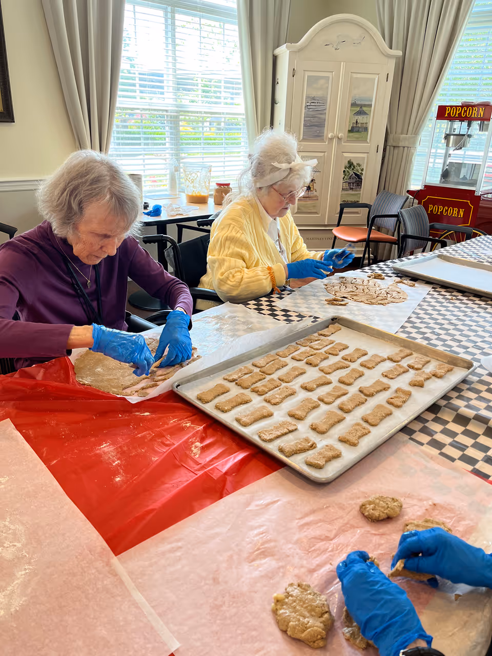 Two elderly women wearing gloves roll and cut cookie dough on a table with baking trays in a bright communal activity room.
