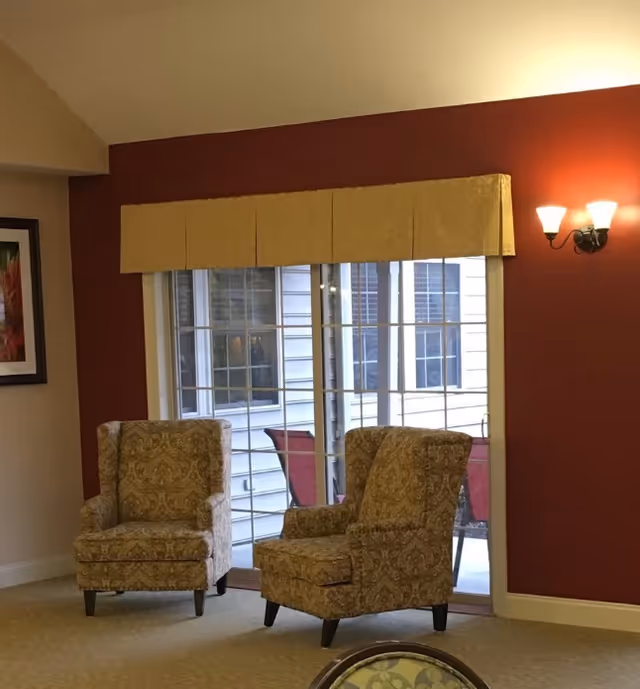 Two patterned armchairs placed near a large window with a yellow valance in a room with red and beige walls. A wall-mounted light fixture with two bulbs is on the red wall, and a framed picture is partially visible on the beige wall.