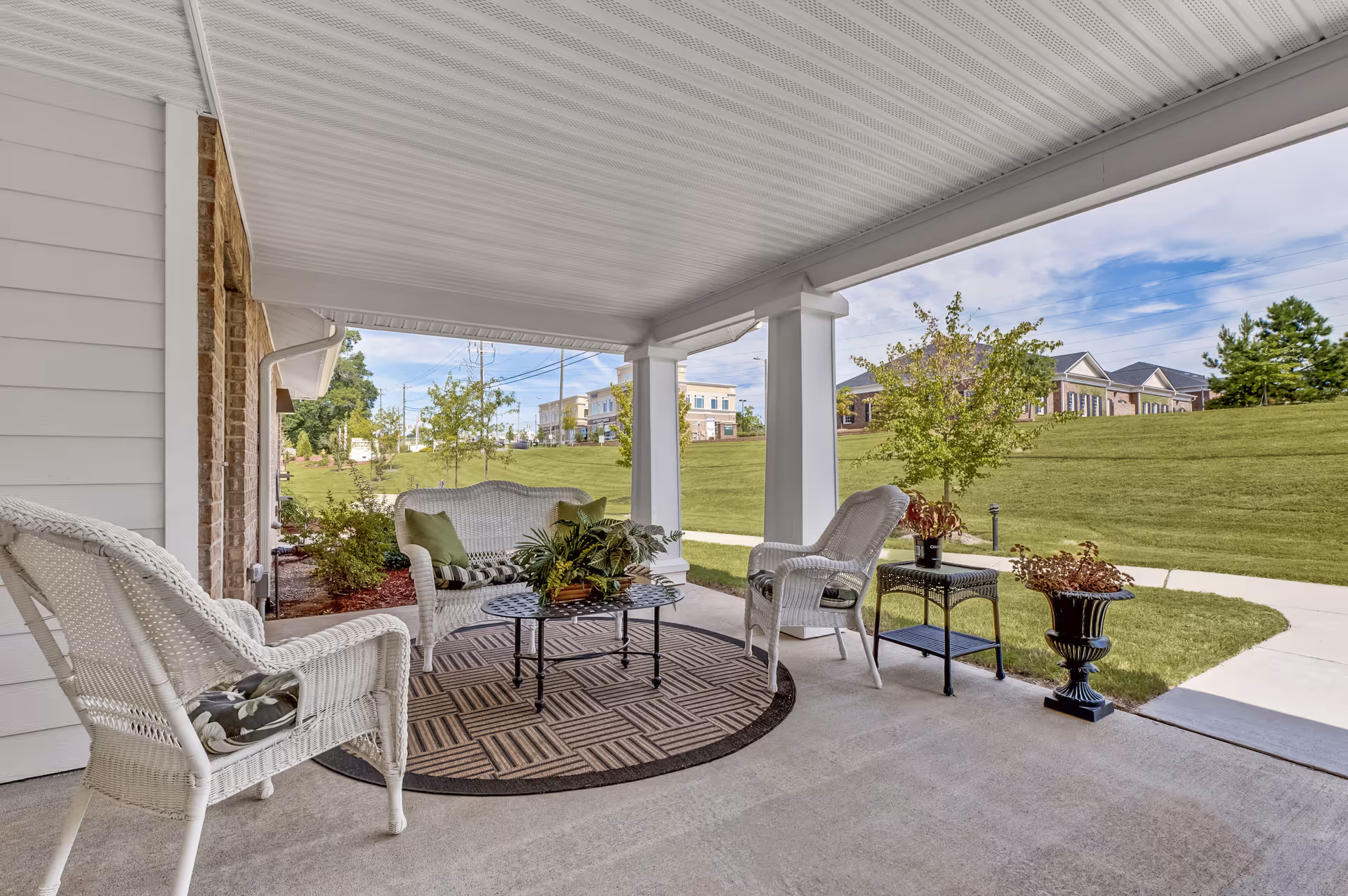 Covered outdoor patio area with white wicker furniture including two chairs and a loveseat with cushions, a round patterned rug, a black metal coffee table with a plant centerpiece, and side tables with potted plants. The patio overlooks a grassy lawn with trees and buildings in the background under a partly cloudy sky.
