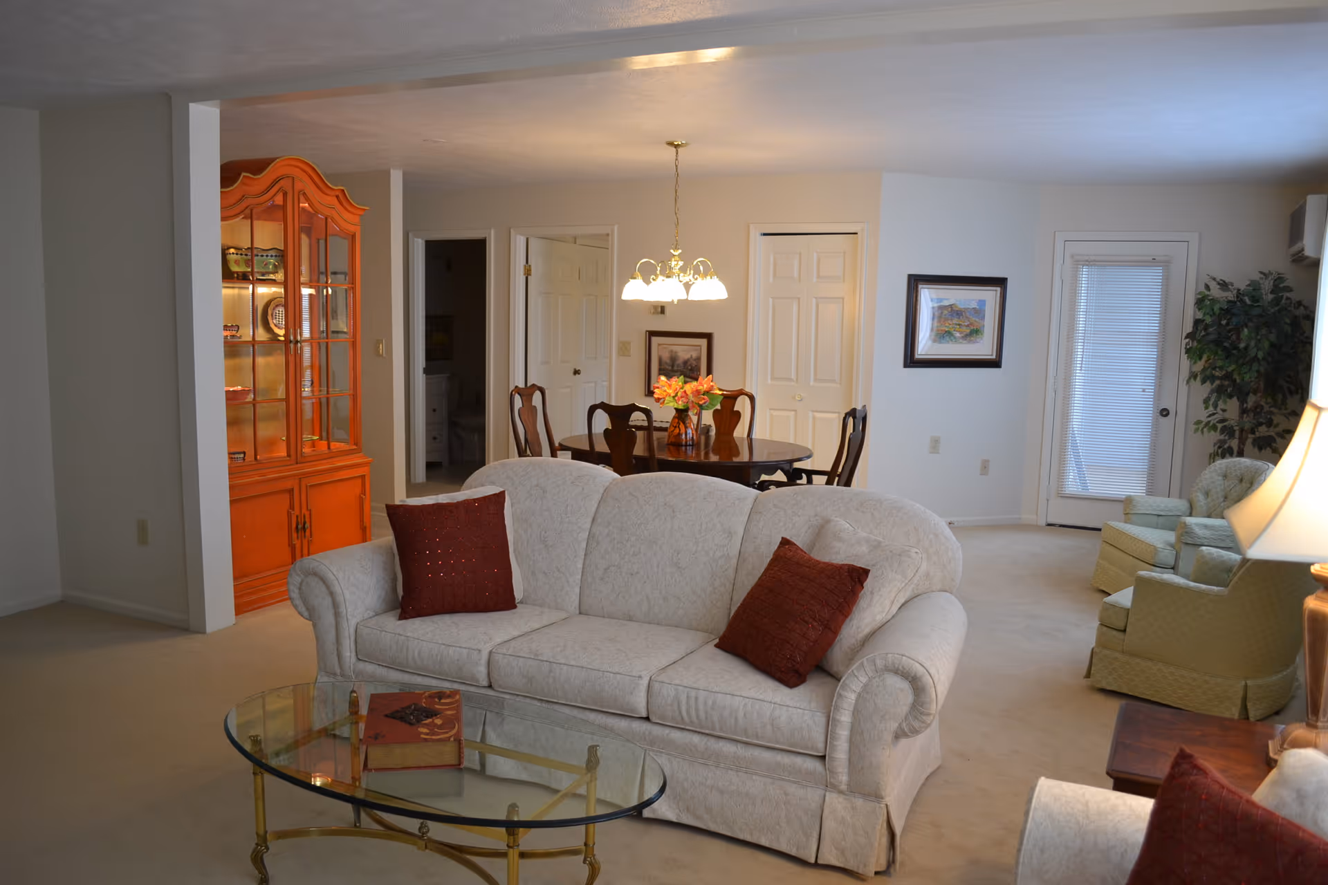 A cozy living room and dining area in a retirement community. The living room features a light-colored sofa with two red pillows, a glass coffee table with a book on it, and two green armchairs near a lamp and a potted plant. The dining area has a wooden table with four chairs and a vase of flowers under a hanging light fixture. A wooden china cabinet with glass doors is visible on the left side, and there are two closed white doors and a framed picture on the wall in the background.
