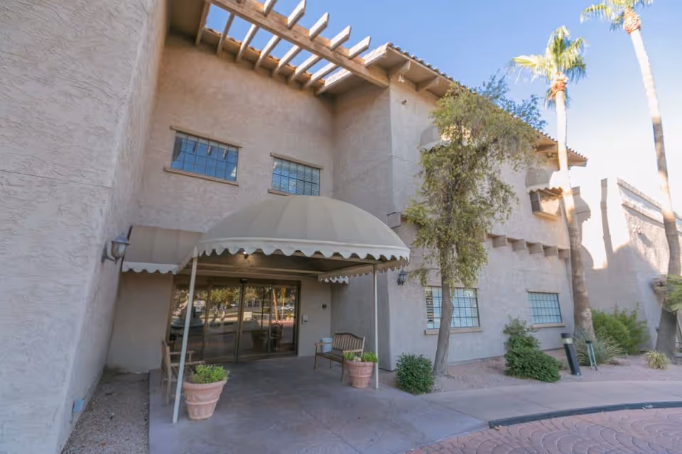 Exterior view of the entrance to a building with a beige stucco facade, a covered awning, glass double doors, potted plants, a bench, and some trees nearby under a clear blue sky.