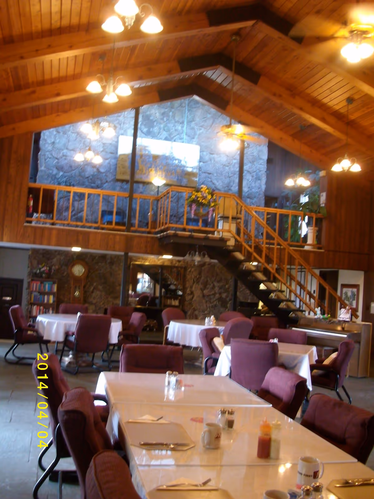 Interior view of a dining area with multiple tables covered with white tablecloths and surrounded by purple upholstered chairs. The room features a high wooden ceiling with exposed beams, several hanging light fixtures, a stone wall, a staircase leading to an upper level with wooden railings, and a grandfather clock near a bookshelf in the background.