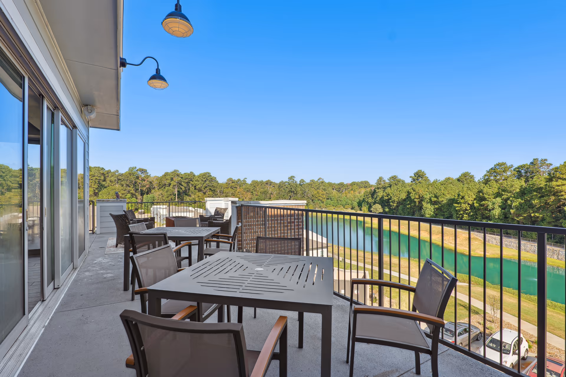 Outdoor balcony area with several tables and chairs overlooking a scenic view of a river or lake and surrounding trees under a clear blue sky.