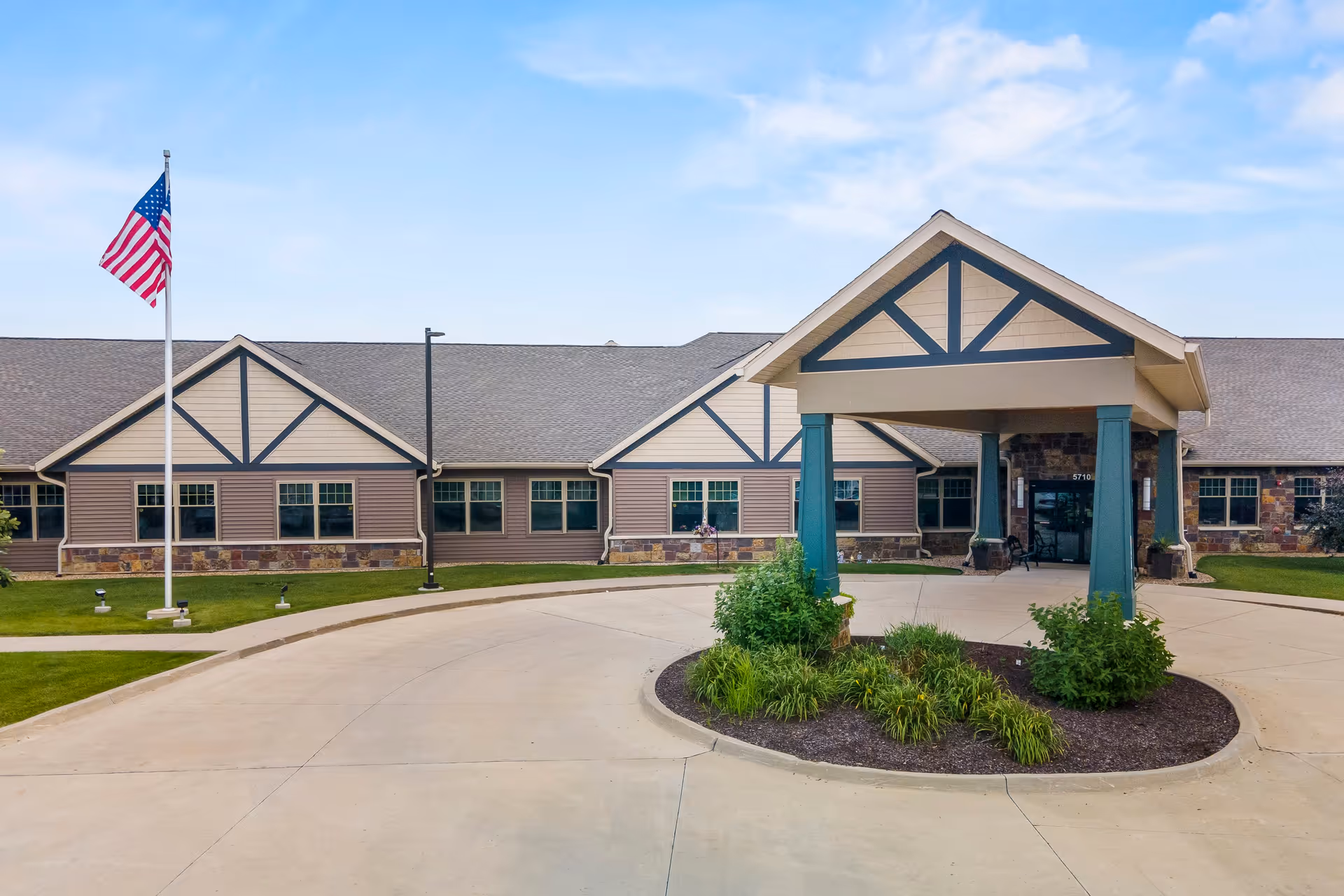 Front exterior view of The Gardens of Cedar Rapids facility showing a single-story building with a covered entrance supported by teal pillars, an American flag on a flagpole, and landscaped greenery around the driveway.