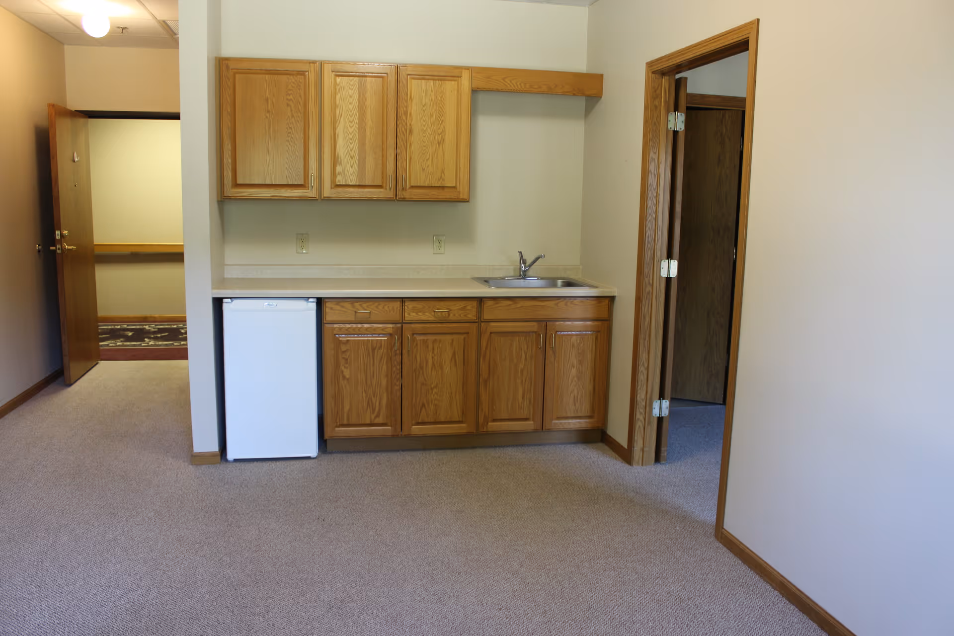 Small kitchenette area with wooden cabinets above and below a countertop, a small white refrigerator, and a stainless steel sink. The room has beige walls and carpeted floor. There is an open doorway to the right and an open door leading to a hallway on the left.