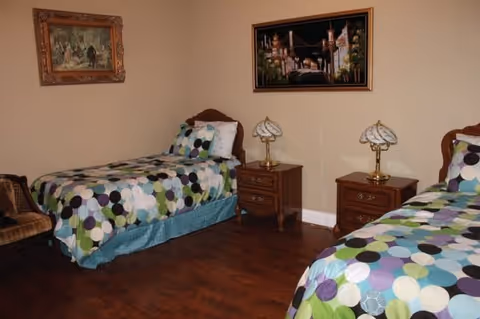 A bedroom with two single beds featuring colorful polka dot bedding. Between the beds are two wooden nightstands, each with a decorative lamp. The room has wooden flooring and beige walls adorned with two framed paintings.