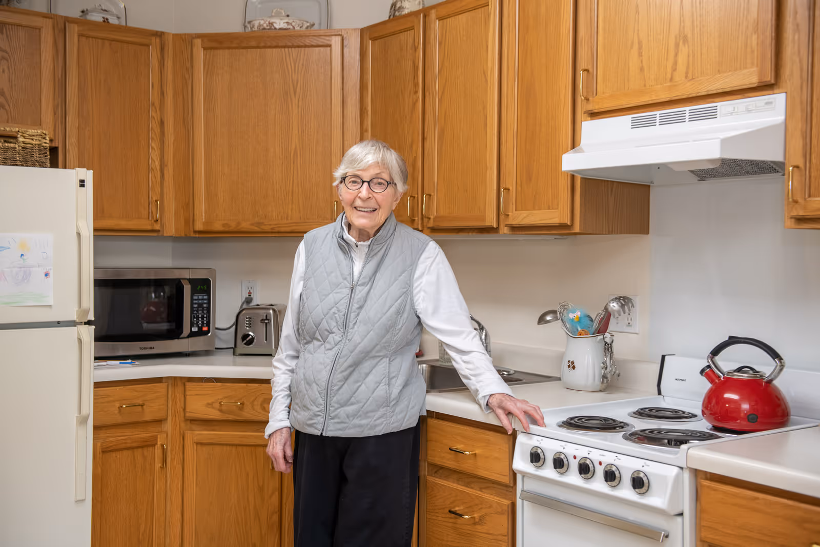 An elderly woman with short gray hair and glasses stands in a kitchen. She is wearing a light gray quilted vest over a white long-sleeve shirt and black pants. The kitchen has wooden cabinets, a white refrigerator with children's drawings attached, a microwave, a toaster, a white stove with a red kettle on it, and various kitchen utensils in a container on the counter.
