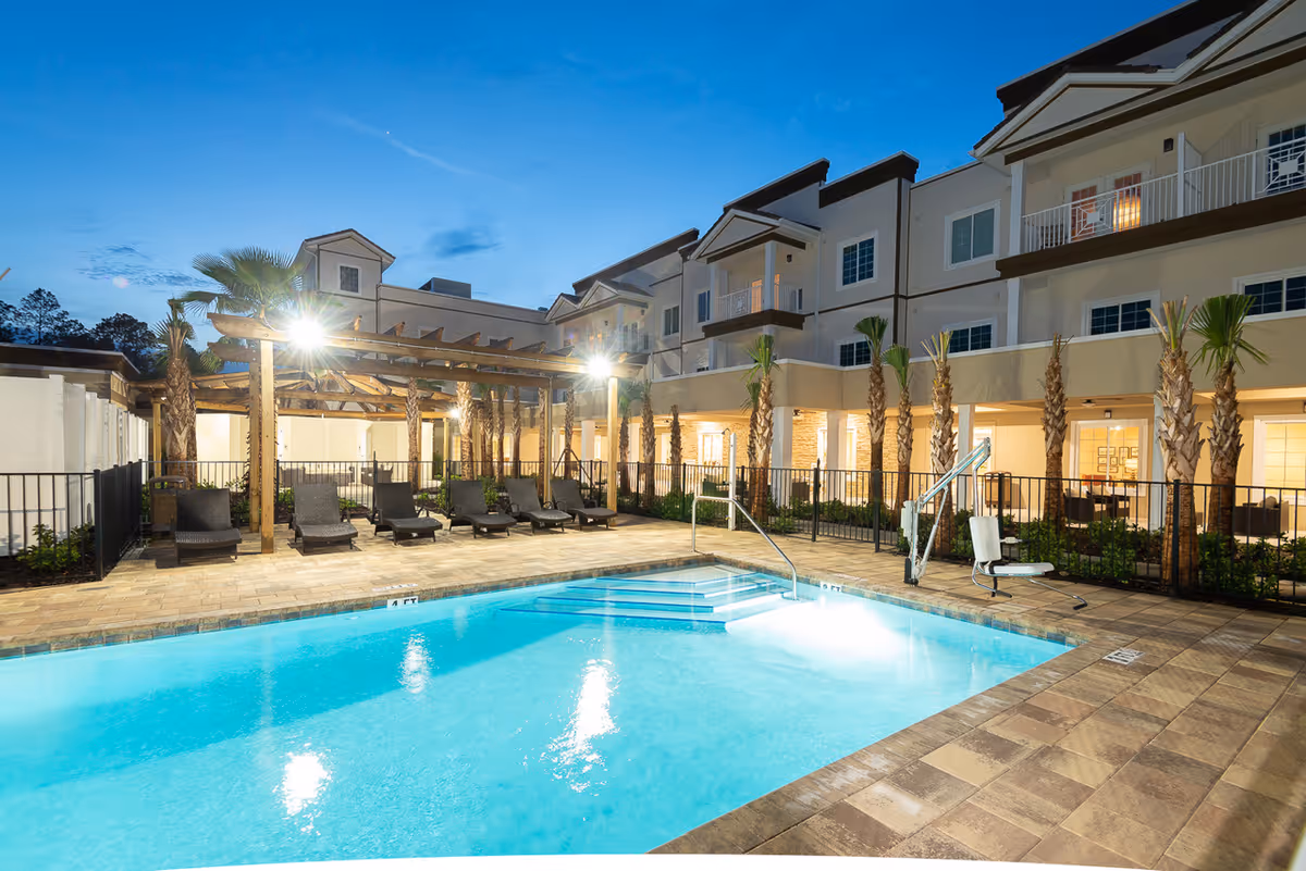 Outdoor swimming pool area at dusk with lounge chairs under a wooden pergola, palm trees, and a multi-story building in the background. The pool has steps and a pool lift for accessibility.