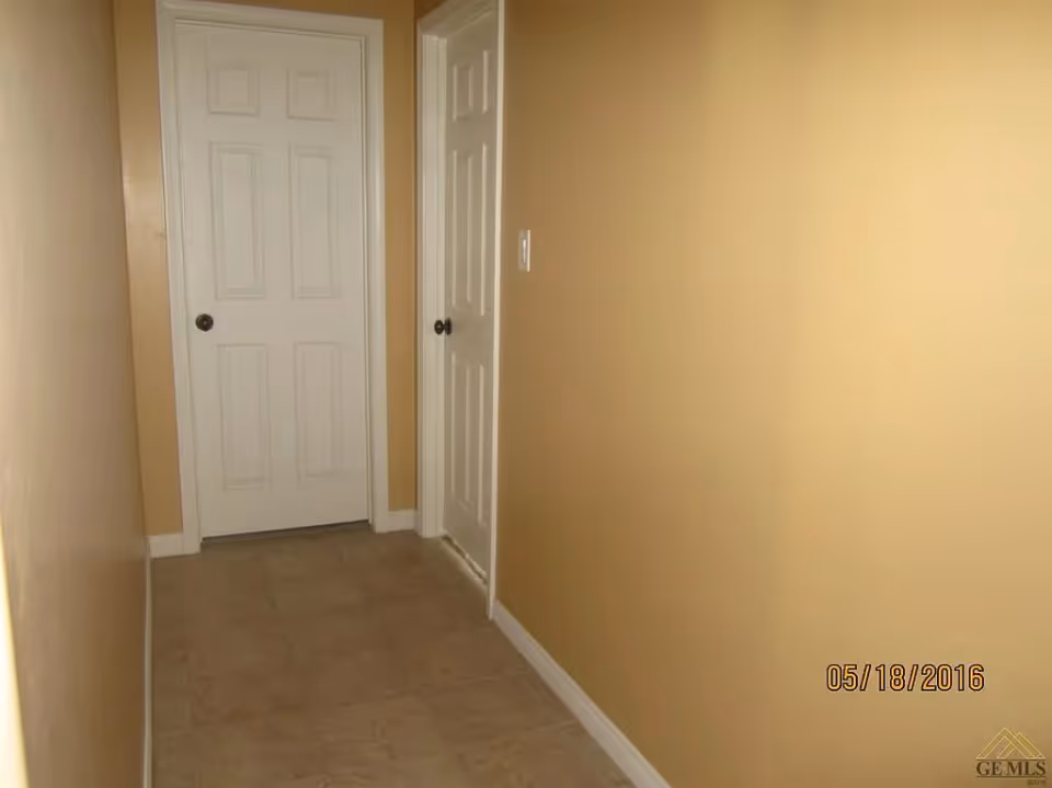 A narrow indoor hallway with beige walls and tiled floor, featuring two white doors with dark round knobs, one at the end of the hallway and one on the right side.