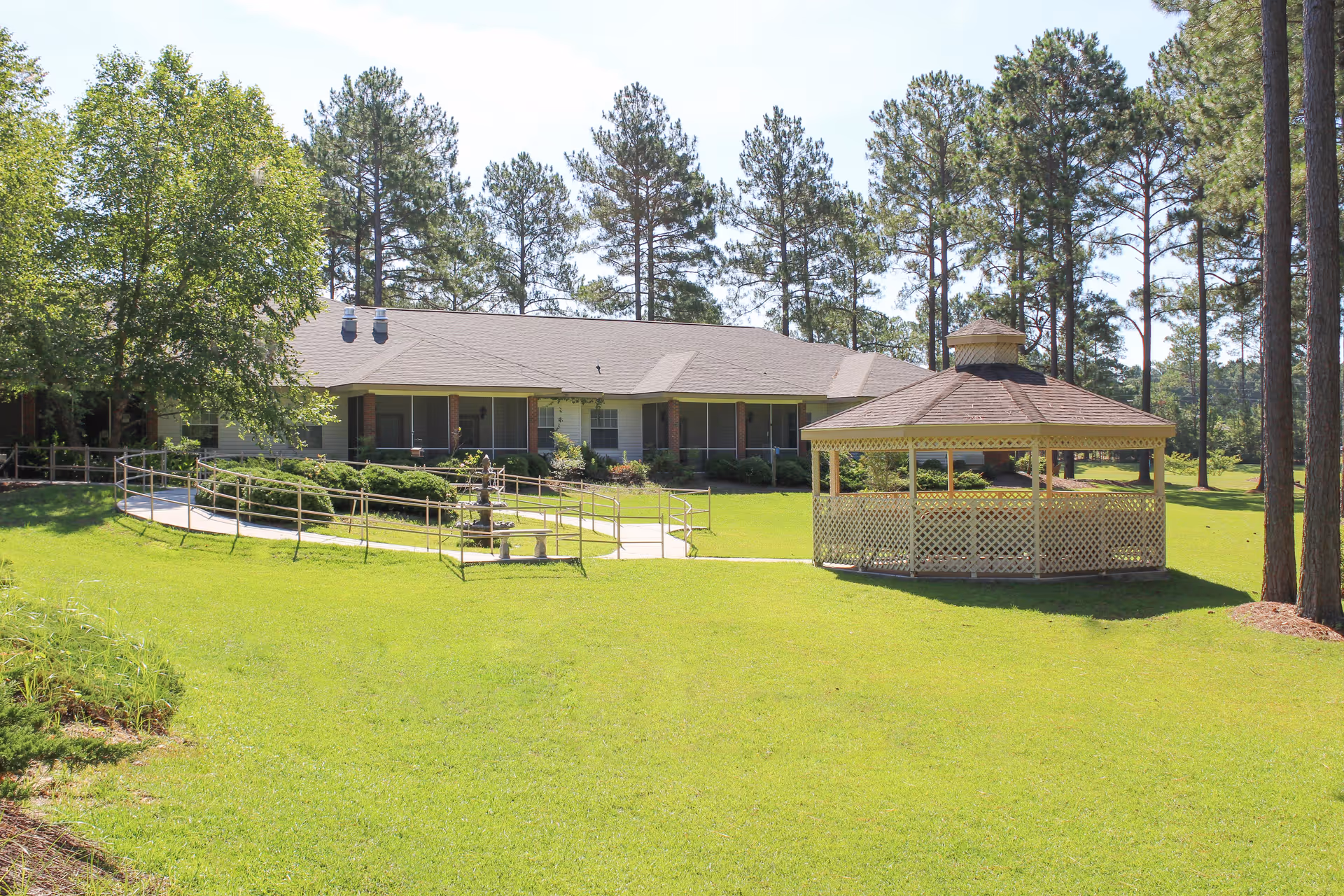 A single-story building with a ramp leading to the entrance, surrounded by green grass and tall pine trees. To the right, there is a wooden gazebo with lattice sides. The sky is clear and sunny.