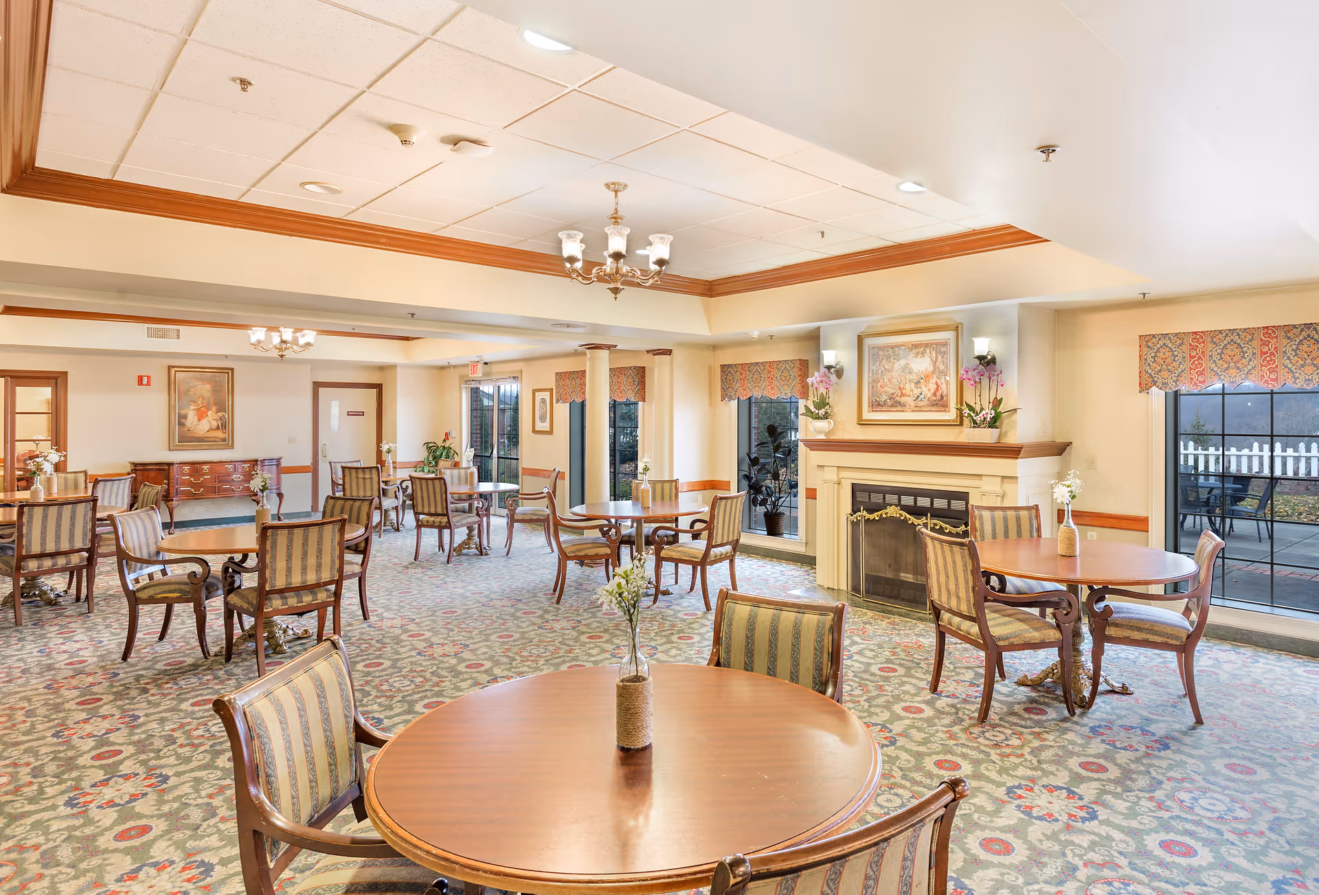 A spacious dining room in a retirement facility with multiple round wooden tables and striped upholstered chairs. The room features a decorative carpet, a fireplace with a painting and flower arrangements above it, large windows with patterned valances, and ceiling light fixtures. There is a door leading outside and a sideboard with a framed picture on the wall.